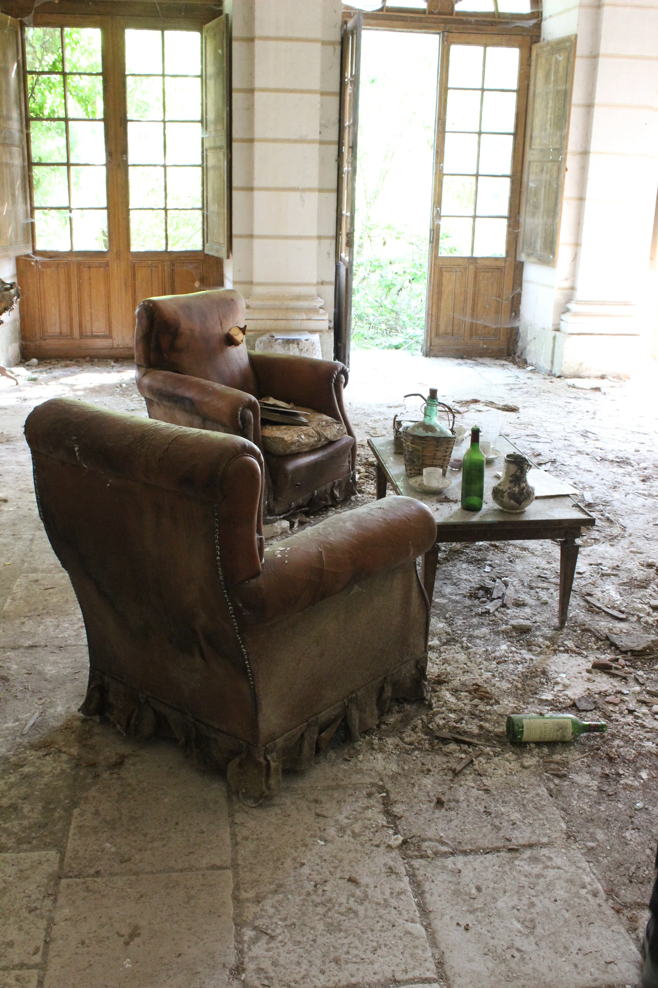 Verfallene, alte Sitze mit einem kleinen Tisch in einem verfallenen Raum mit großen Fenstern und offener Tür, überall Staub und Schmutz, einige leere Flaschen und zerbrochenes Geschirr auf dem Tisch.