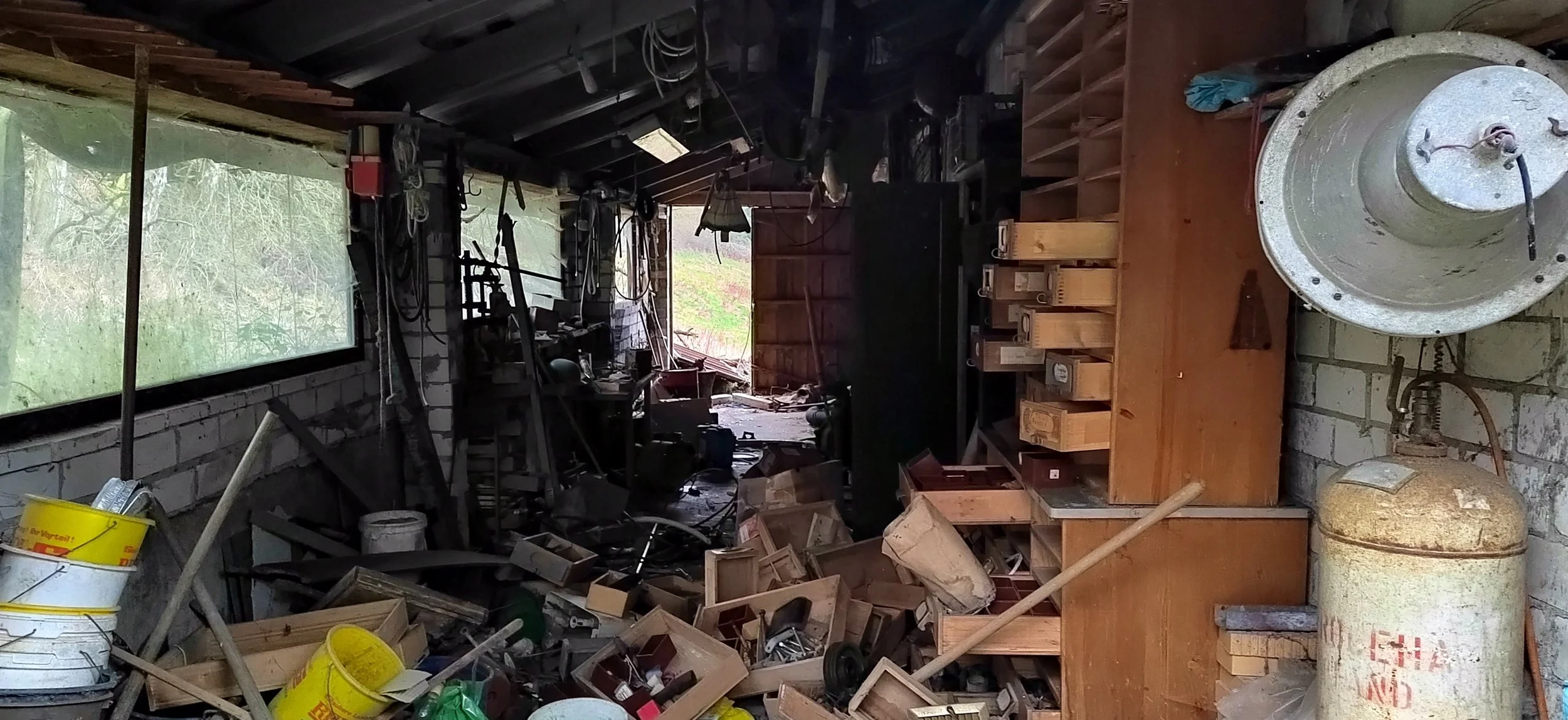 Ein chaotischer, unordentlicher Raum mit Werkzeugen, Holzresten und Verpackungsmaterialien, wahrscheinlich eine Werkstatt oder Garage.