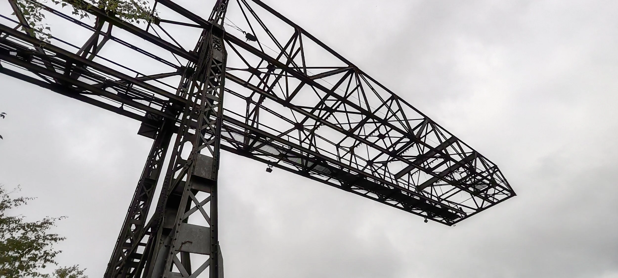 Ein großes, rostfarbenes Eisengerüst einer alten Eisenbahn- oder Hochleitungbrücke gegen einen grauen, bewölkten Himmel.