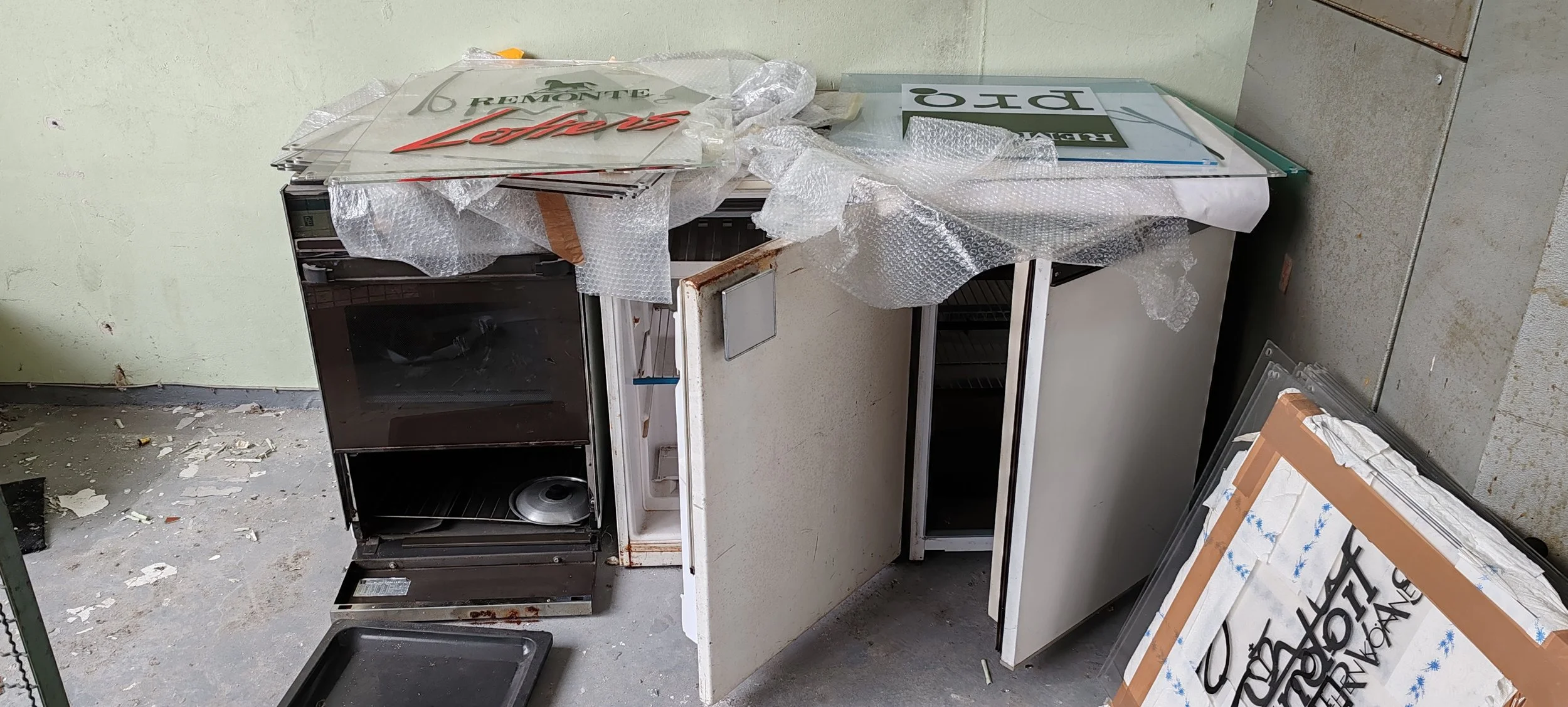 Ein alter, verrosteter Kühlschrank neben einem alten, abgenutzten Thekenbereich. Es liegen Verpackungsmaterialien und beschädigte Schilder auf dem Boden und auf der Theke.