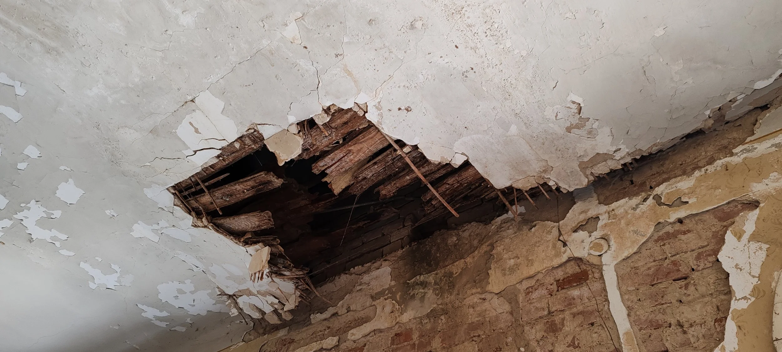 Decke mit großem Loch, abblätternder Farbe und sichtbaren Holzbalken, darüber eine Ziegelwand
