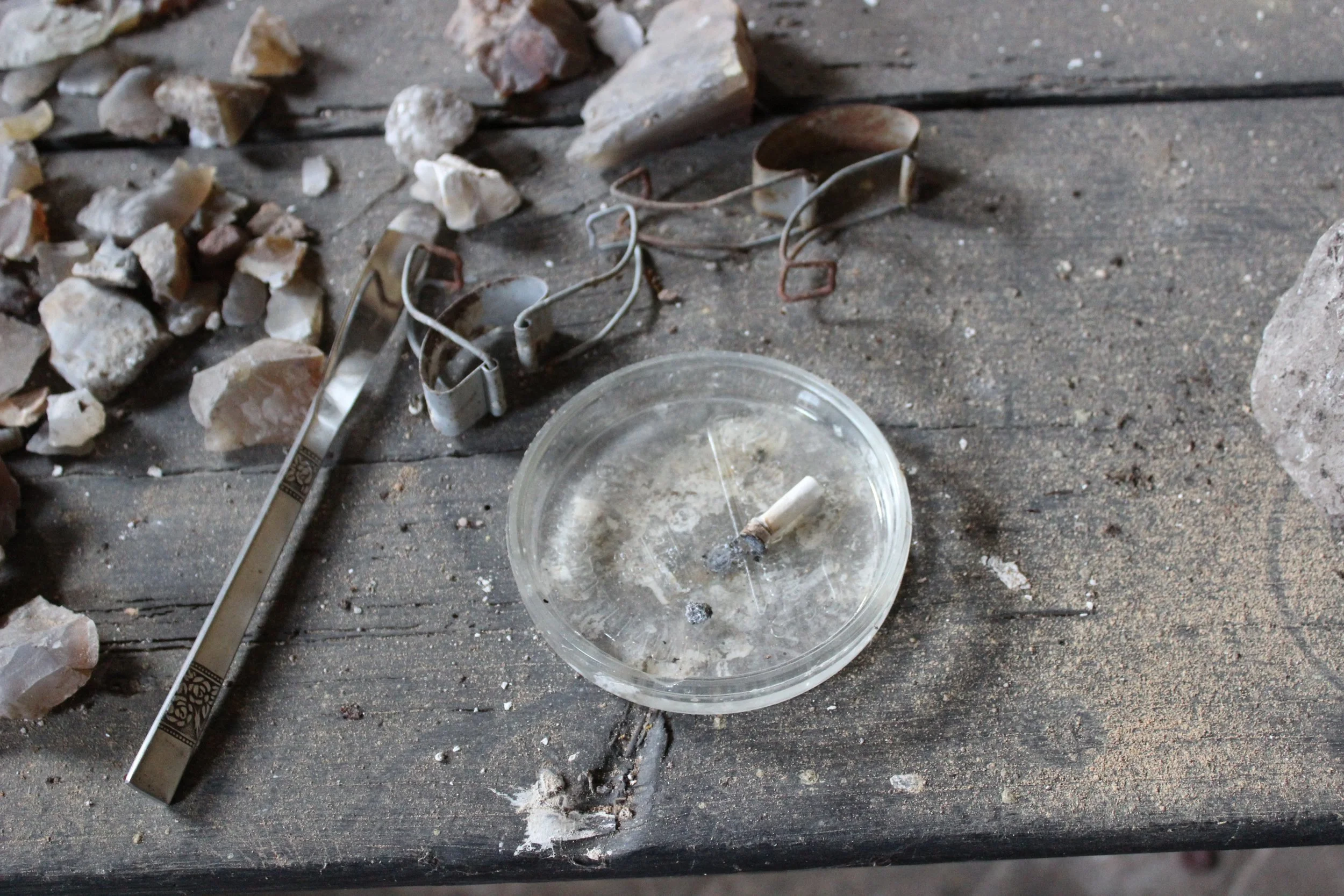 Aschenbecher mit Zigarettenkippe, umgeben von Steinen, rostigen Brillenrahmen, einer Metallleiste und Staub auf einem alten Holztisch.