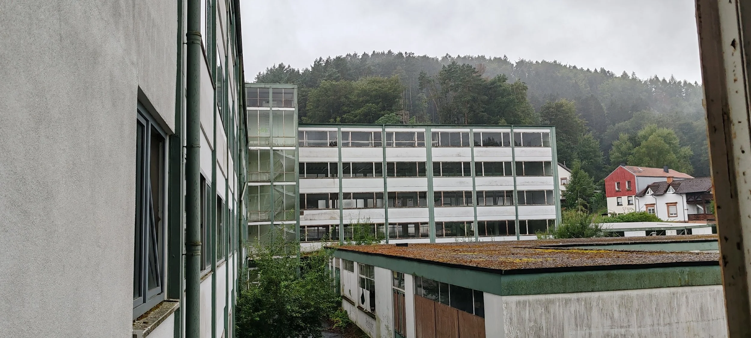 Blick aus einem Fenster auf eine verfallene, mehrstöckige Gebäudefront mit vielen Fenstern, im Hintergrund eine grüne bewaldete Hügelkette, bei leichtem Regen.