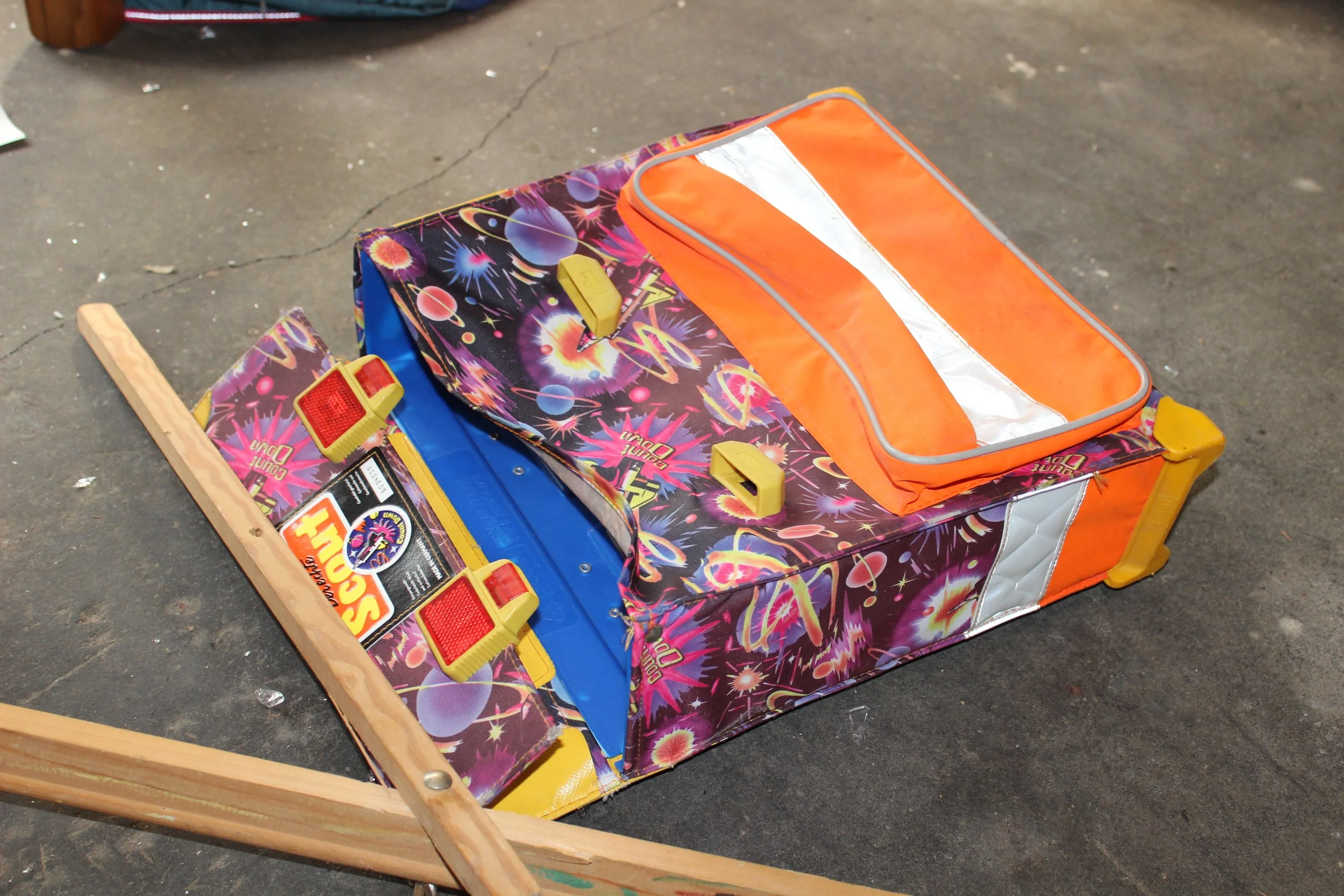 Ein auf dem Boden liegendes Kinderkarussell mit einem bunten, feuerscheinähnlichen Design, orangefarbenem Seitenteil und roten Sicherheitsverschlüssen, auf einem grauen Boden.