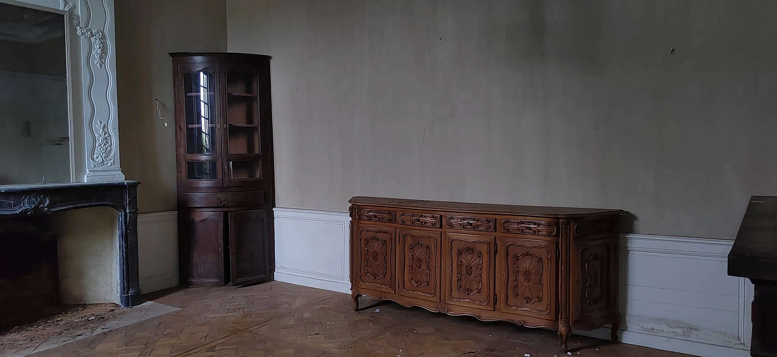 Ein leerer Raum mit einer traditionellen Einrichtung, darunter ein dunkler Holzschrank, eine elegante Holzkommode und ein großer Spiegel mit verzierter weiße Rahmen. Der Boden besteht aus Parkett, der Raum wirkt etwas unordentlich und nicht renoviert