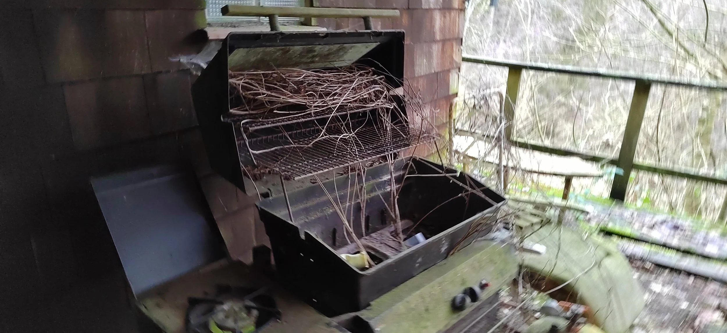 Verrosteter, vernachlässigter Grill mit Spinnenweben auf einer Terrasse, im Hintergrund ein Holzzaun und eine gläserne Scheibe.
