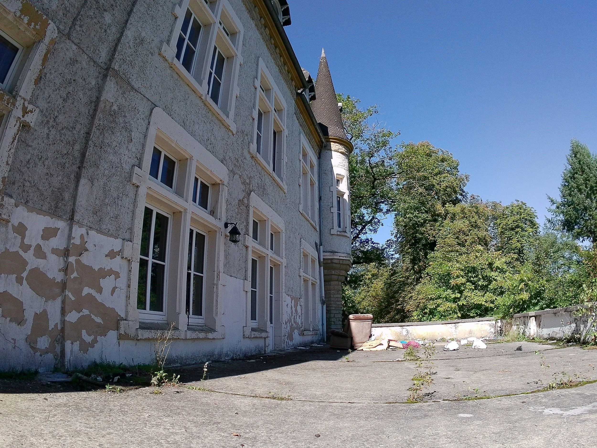 Verfallenes, altes Gebäude mit schäbiger Fassade, mehrere Fenster, ein kleiner erker und ein Turm mit spitzem Dach, daneben einige Bäume und ein blauer Himmel.