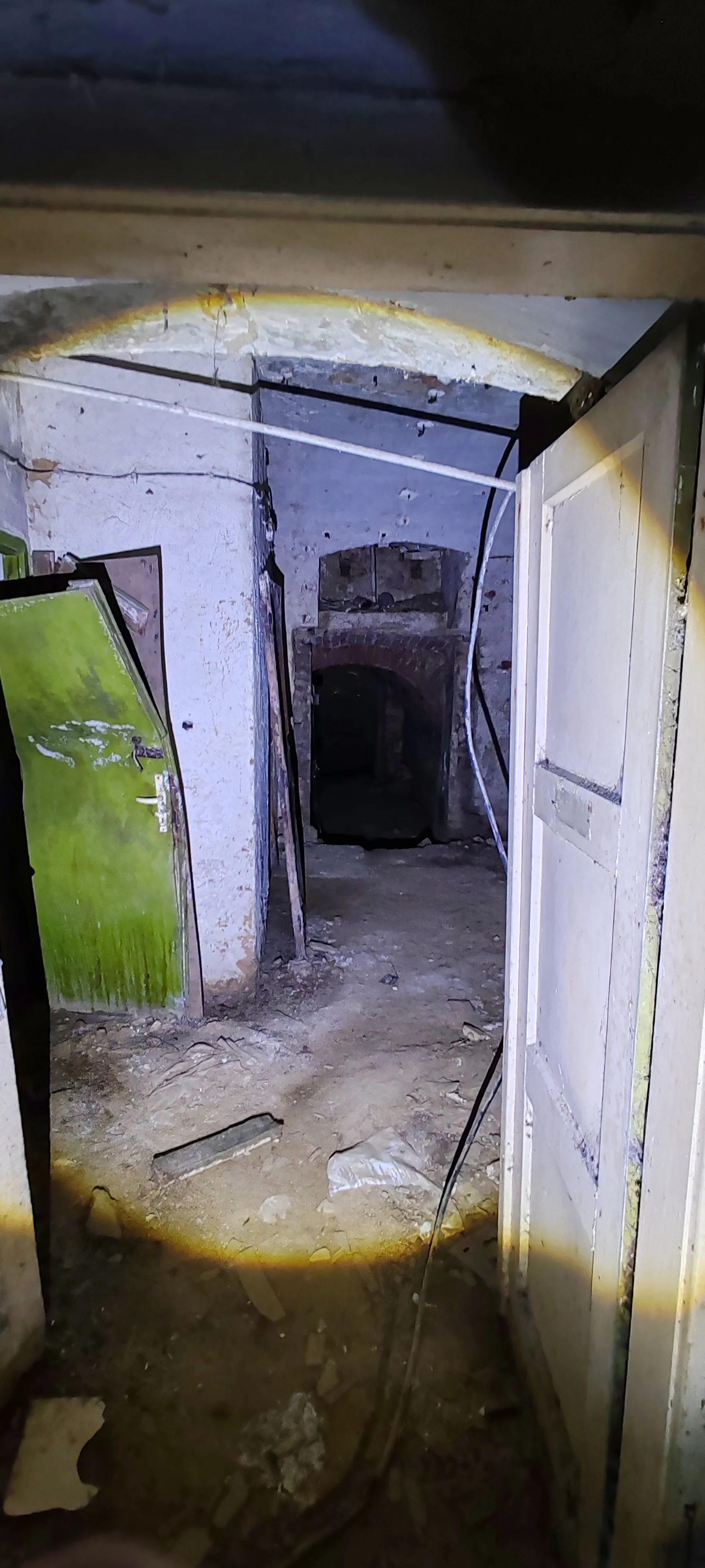 Ein dunkler, verlassener Kellerraum mit Schmutz und Staub auf dem Boden, einem alten Ofen oder Kamin in der Mitte und alten Türen an den Seiten.