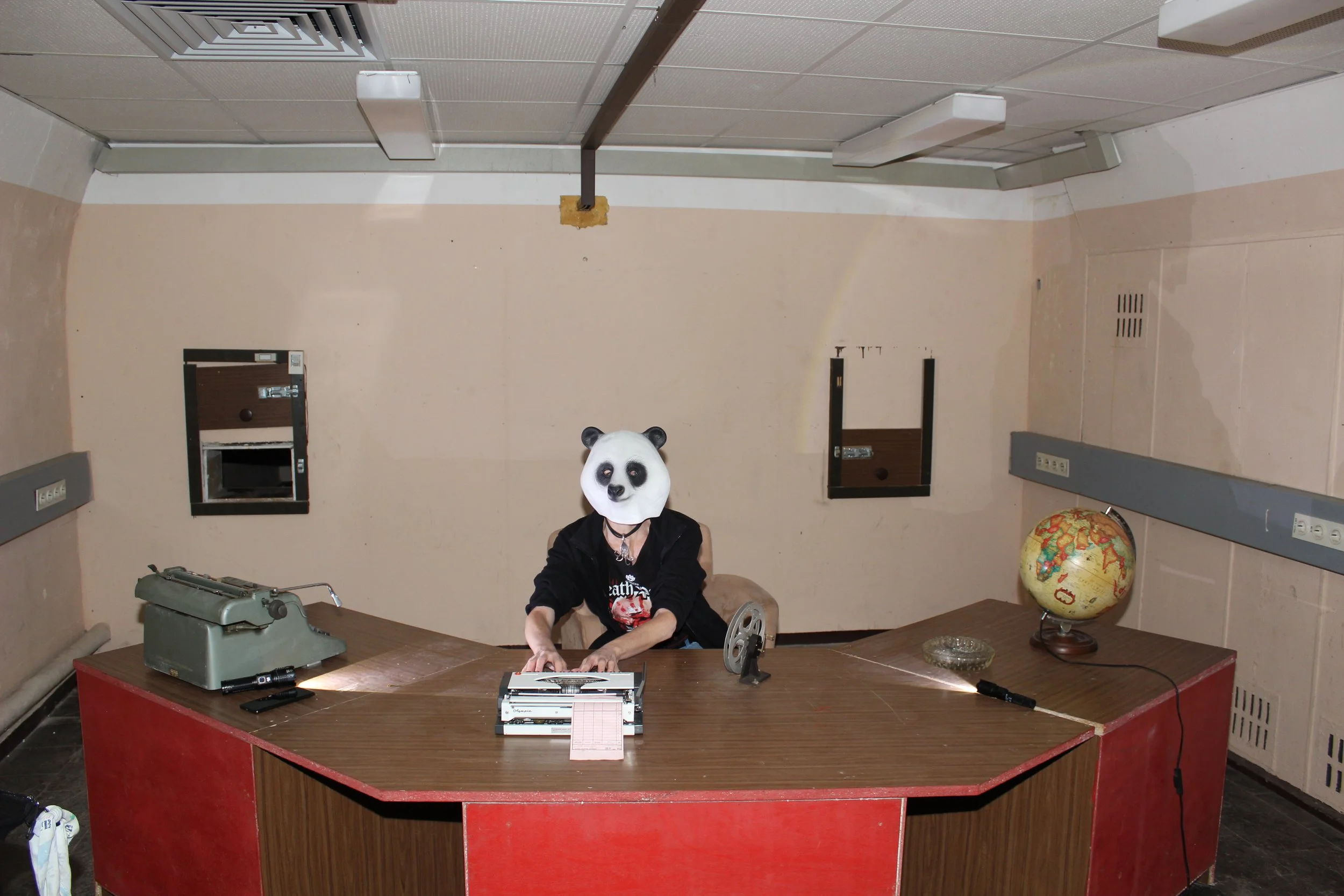 Ein Raum mit einem Schreibtisch, auf dem ein Computer und eine alte Schreibmaschine stehen. Eine Person mit Panda-Maske sitzt am Tisch. Es gibt eine Weltkugel auf dem Tisch und eine Wand mit mehreren Steckdosen.