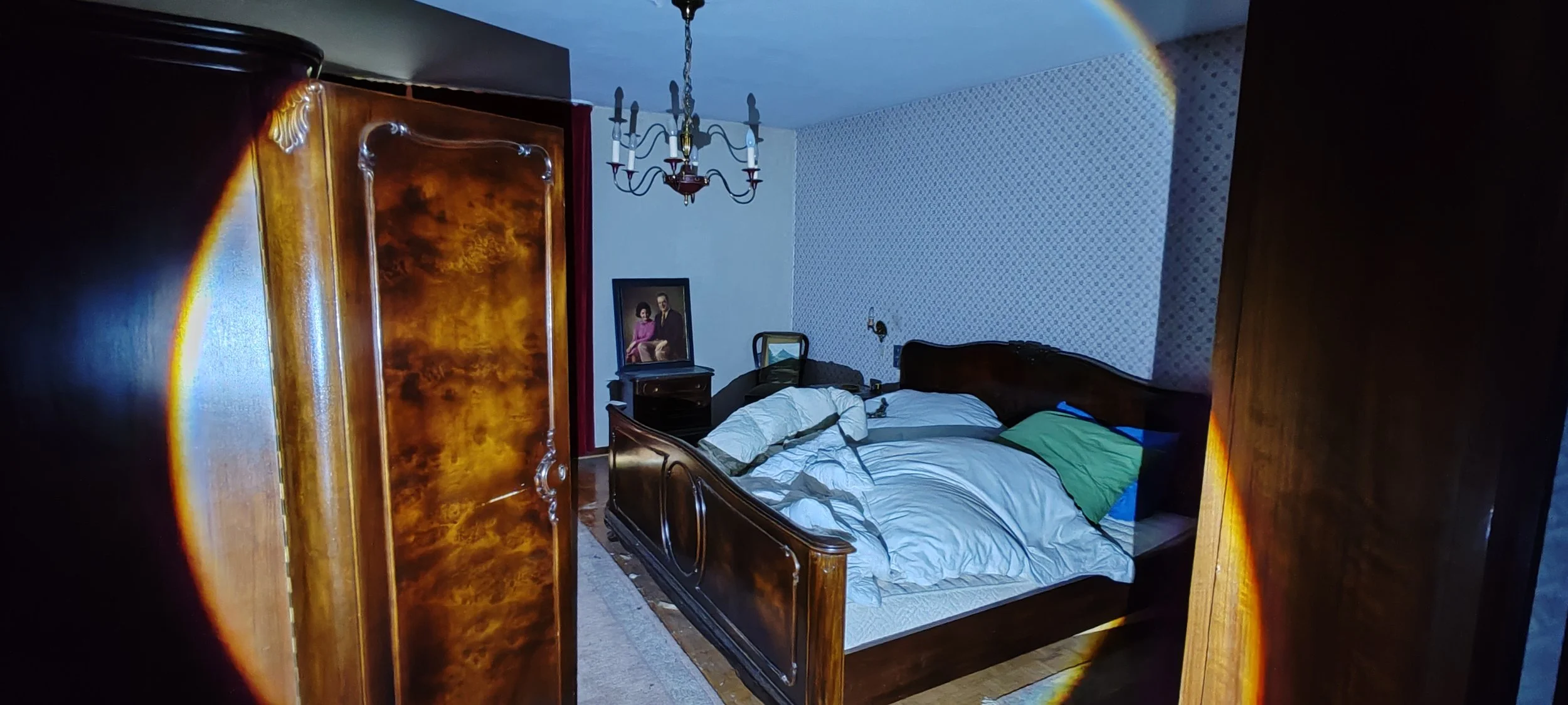 Ein unordentliches Schlafzimmer mit einem großen, dunklen Holzbett, das unaufgeräumt ist, und einem Kleiderschrank, der die Sicht versperrt. Im Hintergrund hängt ein Kronleuchter von der Decke, und auf einem kleinen Nachttisch steht ein Portrait eine