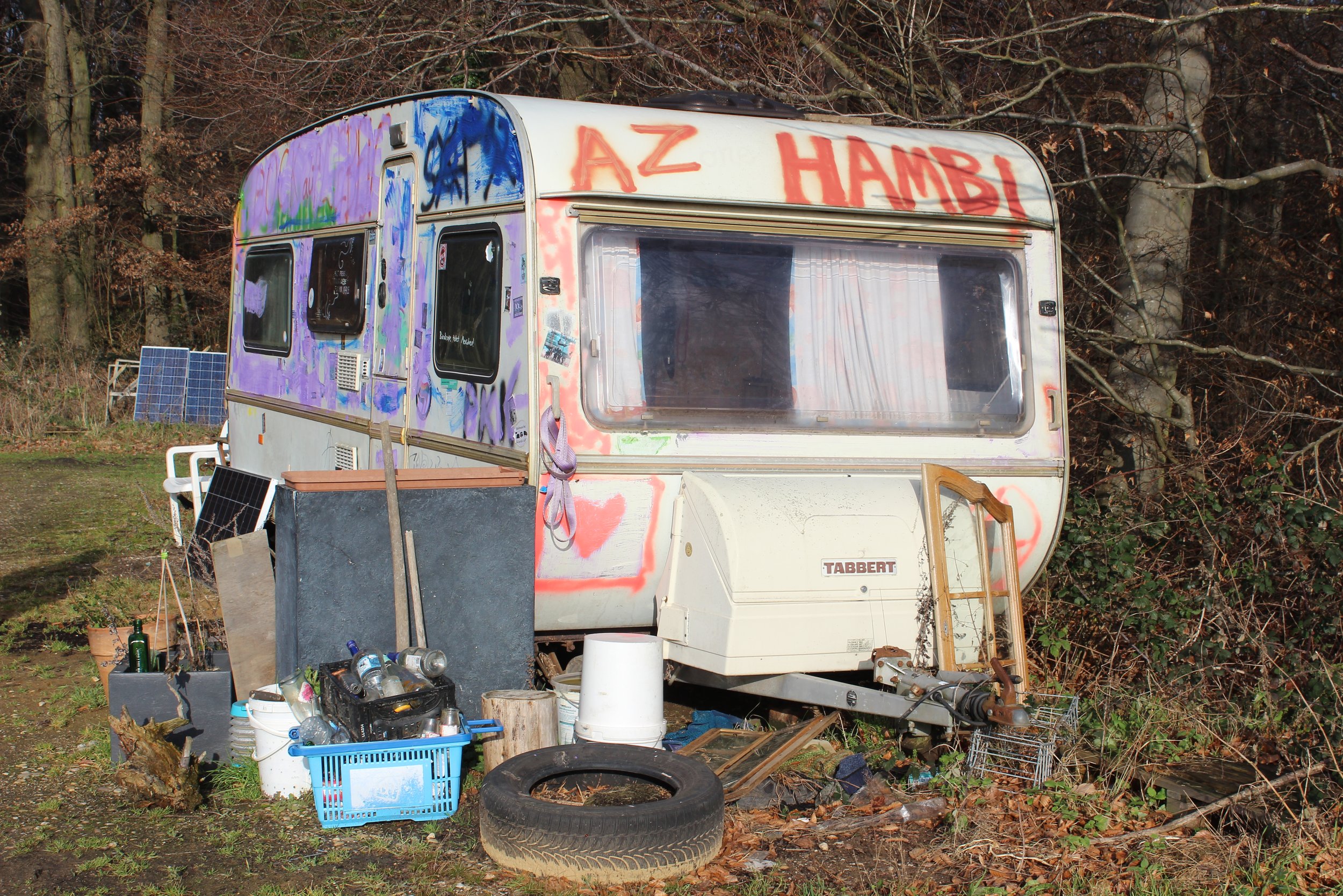 Ein veralteter Wohnwagen mit Graffiti und japanischem Schriftzug vor einem Wald mit Bäumen ohne Blätter, umgeben von Müll und alten Gegenständen.