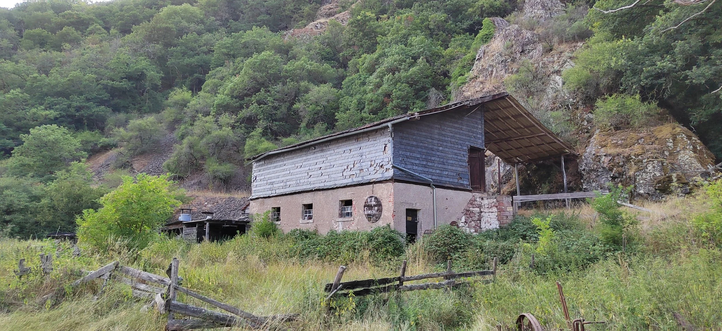 Ein altes Haus in einer ländlichen Umgebung, umgeben von hohem Gras, Bäumen und Felsen im Hintergrund, mit einer Holz- und Zementstruktur, die teils verfallen ist.