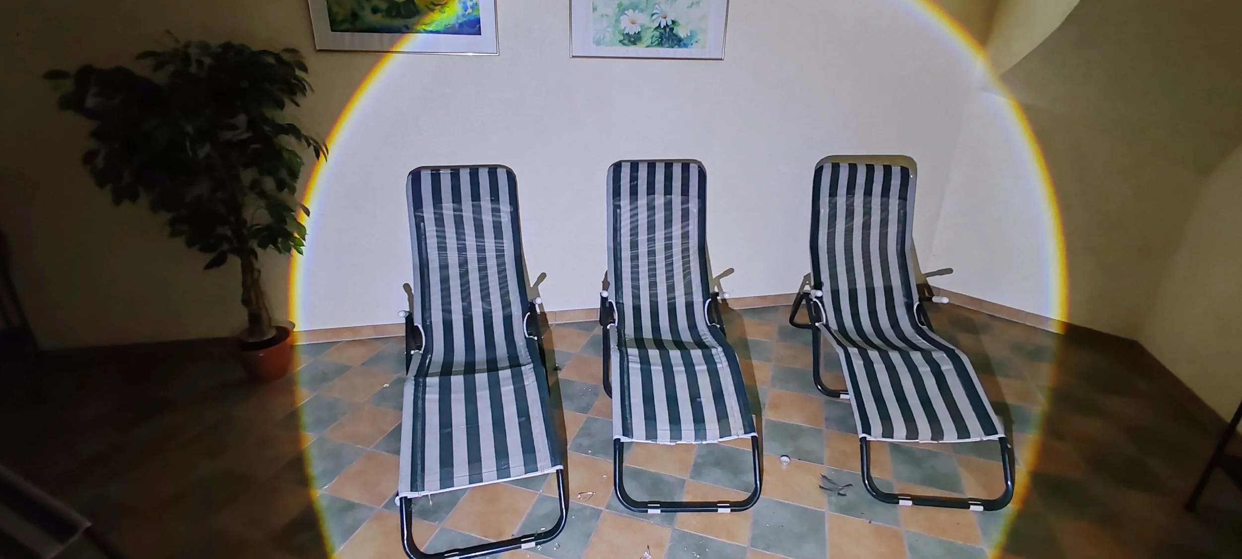 Drei Strandliegen mit gestreiftem Stoff in einem Raum, beleuchtet durch einen Regenbogen-Effekt, mit einem Topfpflanzen und zwei gemalten Bildern an der Wand im Hintergrund.
