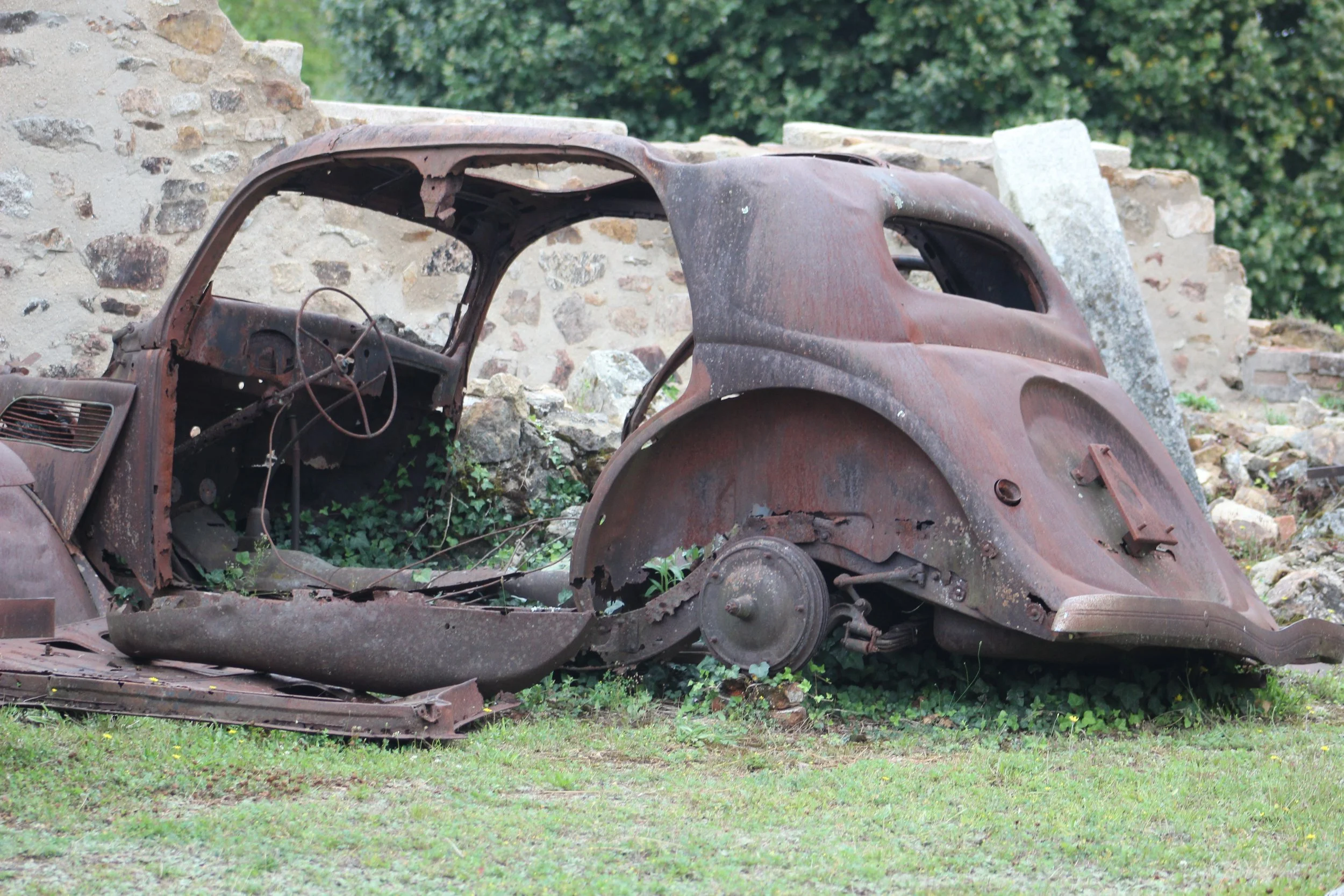 Verrostetes altes Automobilwrack auf Wiese neben zerfallener Steinmauer, Umrisse eines alten Autos, kaum erkennbar, überwuchert mit Grünpflanzen.