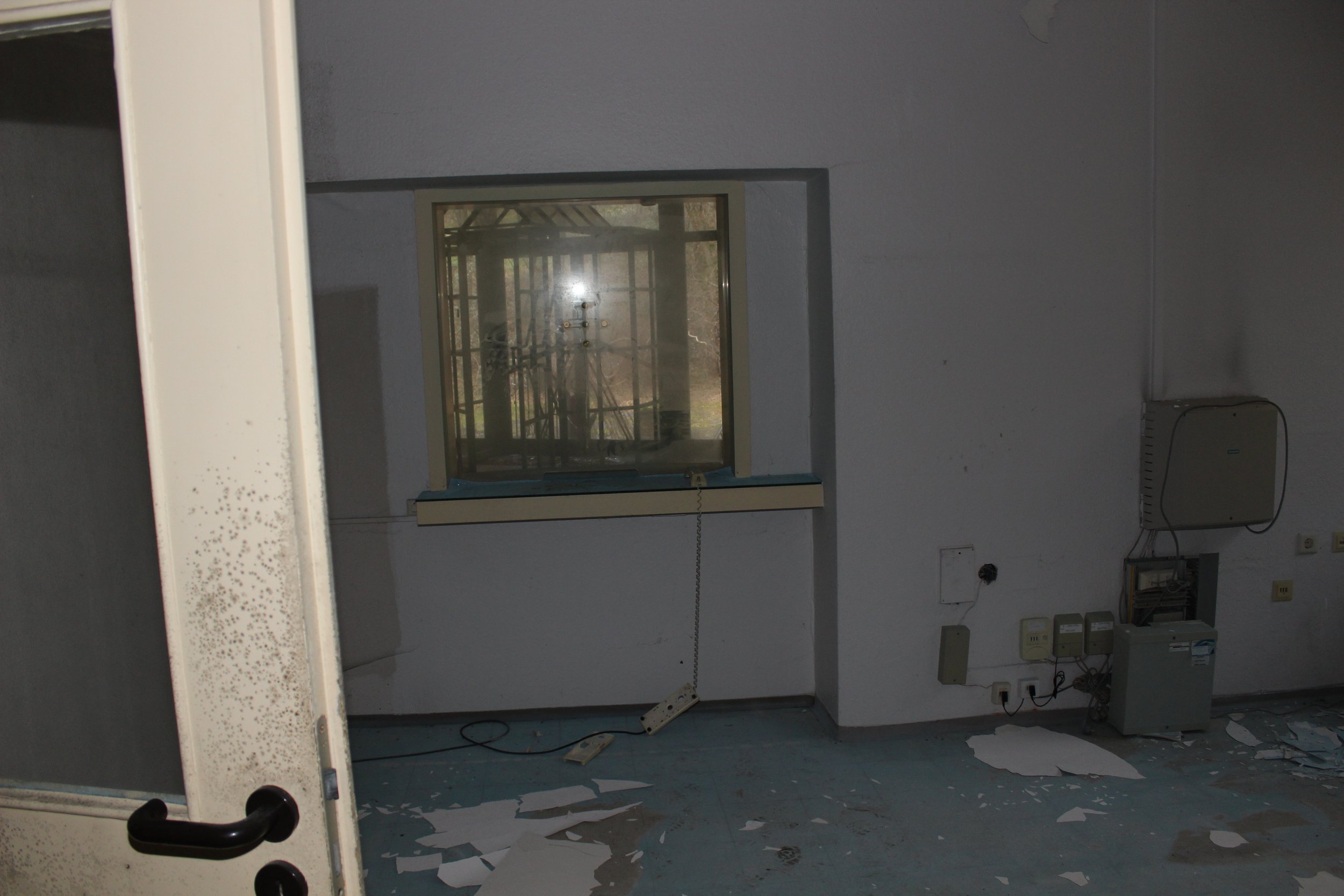 Ein leeres, ungepflegtes Zimmer mit verschmutztem Boden, einem Fenster, einem alten Heizkörper und verschiedenen Kabeln und Schaltern an der Wand.