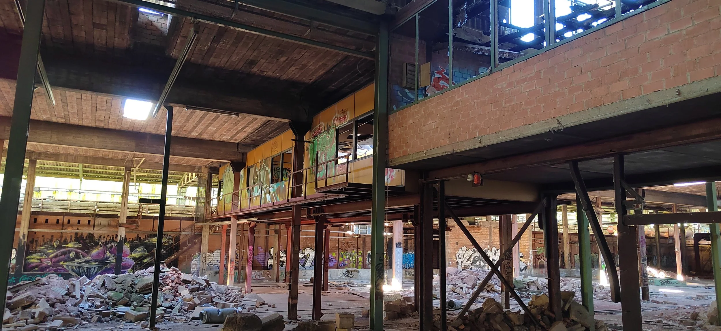 Innenraum eines verfallenden Gebäudes mit Graffiti an den Wänden, Trümmern und Baugerüsten, viel Sonnenlicht durch große offene Flächen.
