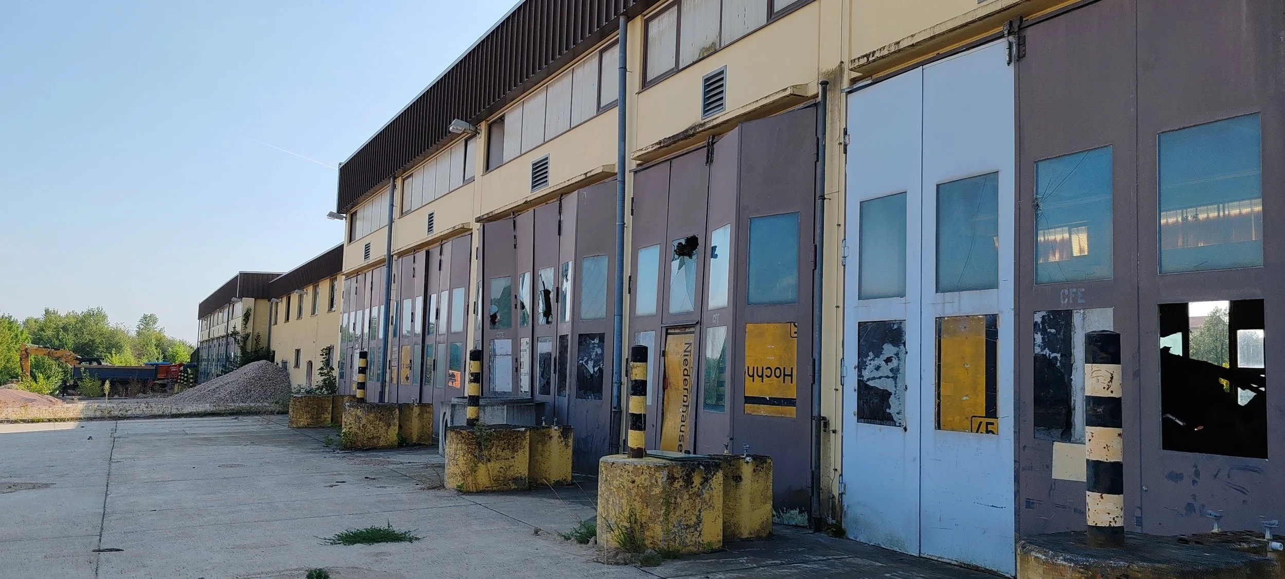 Ein verfallenes, verlassenes Gebäude mit zerbrochenen Fenstern und verrosteten Metallen. Im Vordergrund eine leere Fläche mit einigen Moosbewachsenen Betonblöcken und Müll, im Hintergrund Bäume und Baugeräte.