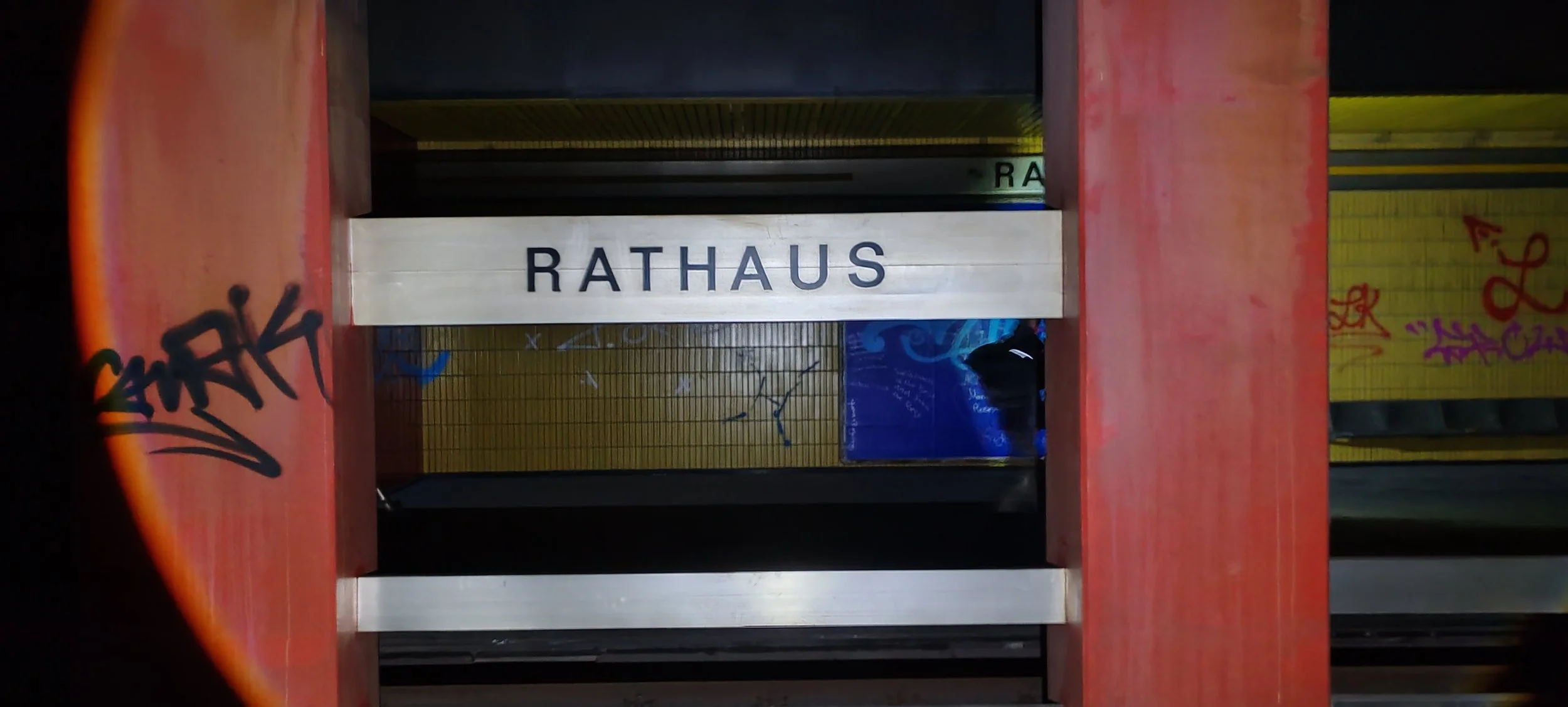 Ein Verkehrsschild mit der Aufschrift 'RATHAUS' in einer Bahnhofshalle mit grauen Wänden, buntem Graffiti und gelben Fliesen im Hintergrund.