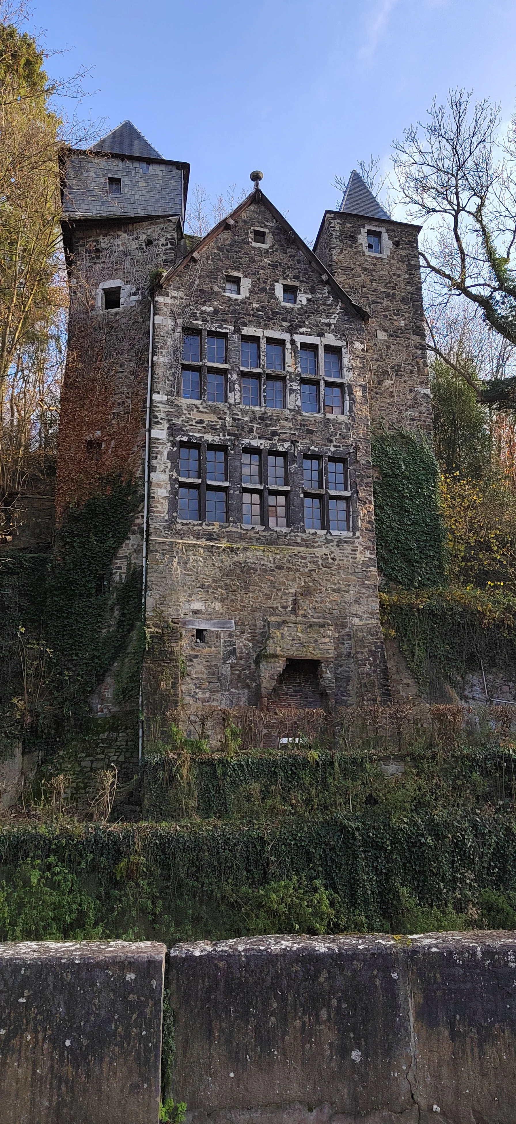 Ein mittelalterlicher Turm aus Stein mit mehreren kleinen Fenstern, umgeben von Bäumen und bewachsen mit Efeu, bei Tageslicht.