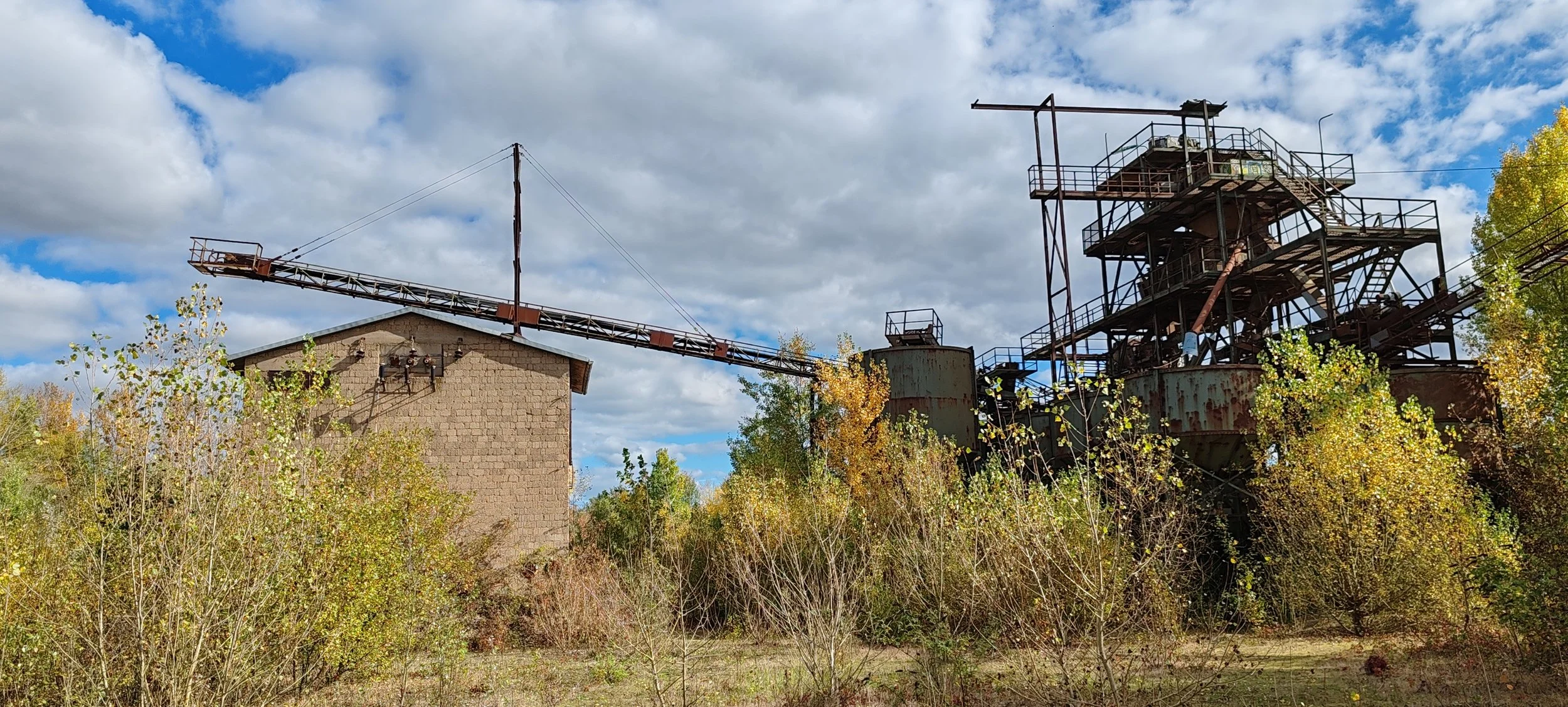 Verlassene, rostige Industrieanlage mit Steigungen und einem langen Förderband, umgeben von Büschen und Bäumen, unter einem bewölkten Himmel.
