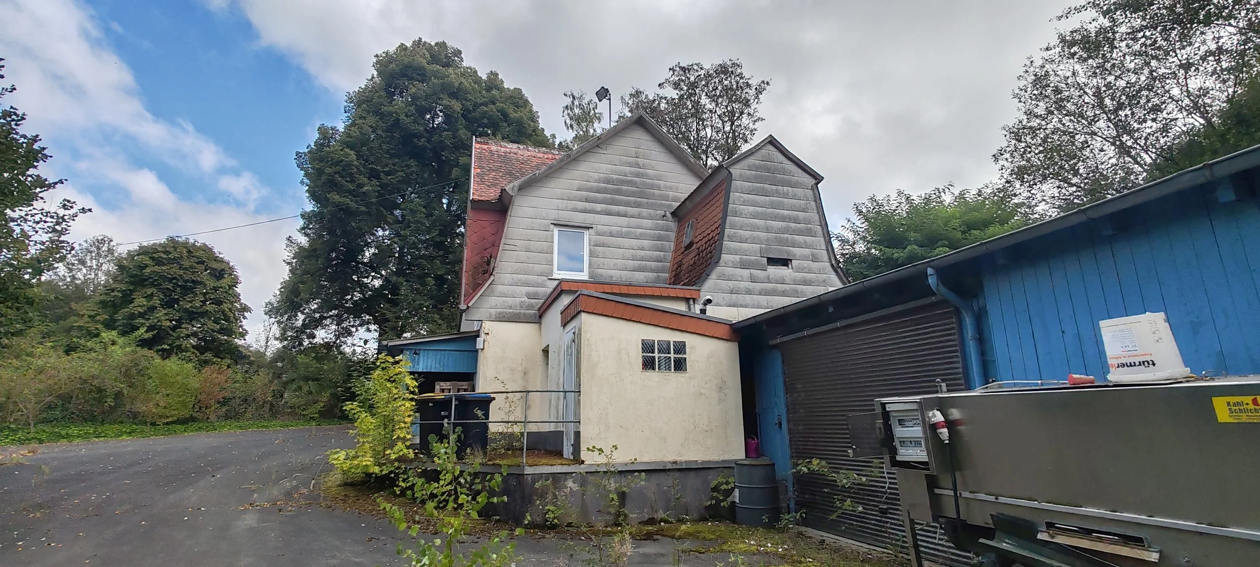 Ein zweistöckiges Haus mit Dachnasen, umgeben von Bäumen, und einem blauen Gebäude mit Schiebetor auf der rechten Seite. Es gibt ein kleines Fenster mit Gitter und eine Regenrinne. Im Vordergrund eine asphaltierte Fläche mit einigen Pflanzen.
