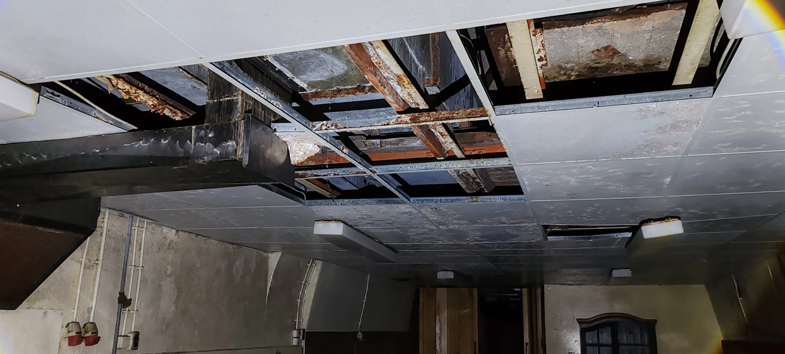 Decke mit beschädigten und abmontierten Deckenplatten, sichtbare Metall- und Holzträger, Putz und Veralterung.