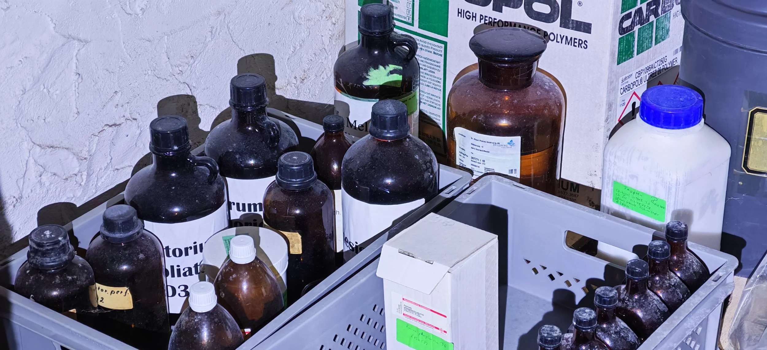 Verschiedene dunkle und helle Chemikalienflaschen in einer Kiste, einige mit weißen Labels, in einer Lagerumgebung.