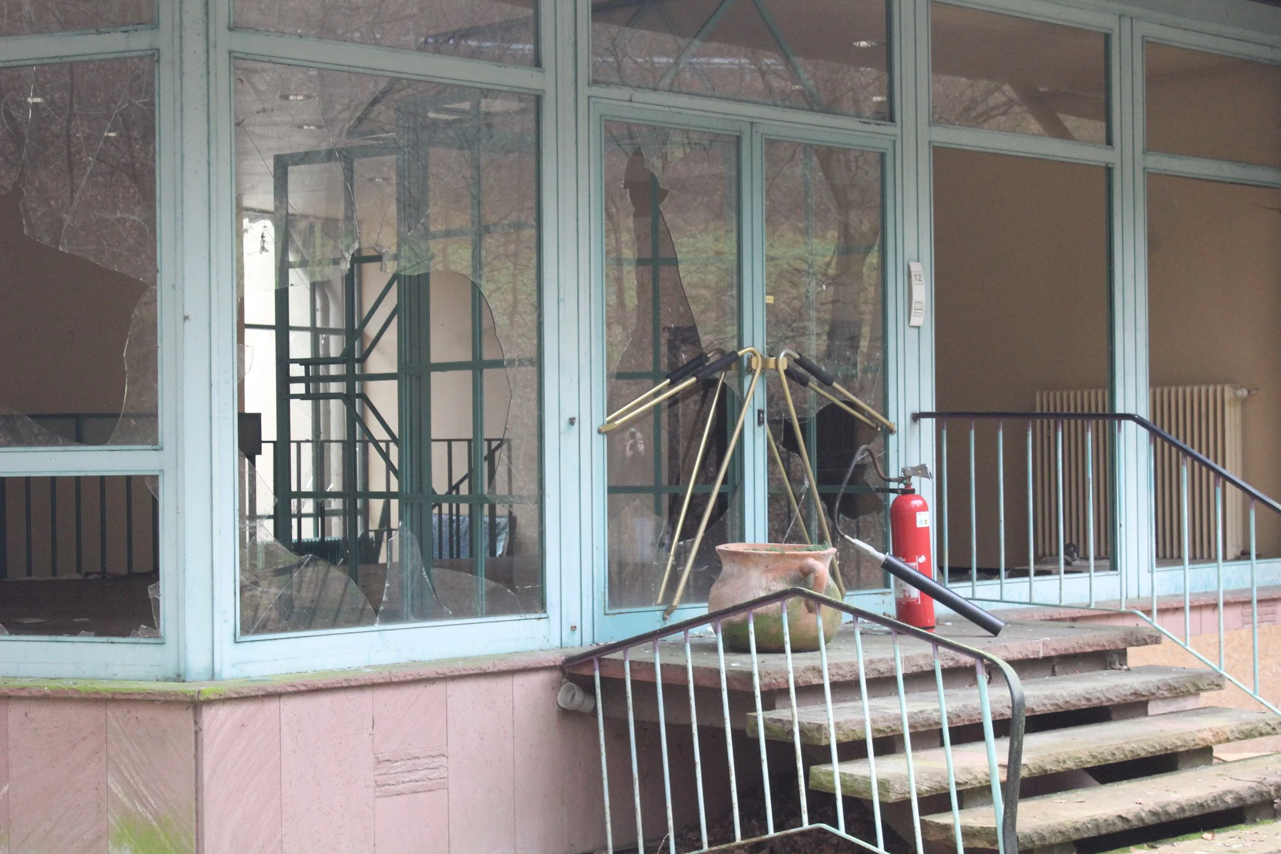 Ein eingestürztes Glasgebäude mit zerbrochenen Fensterscheiben, ein Feuerlöscher, ein alter Keramiktopf und ein Einkaufswagen im Inneren.