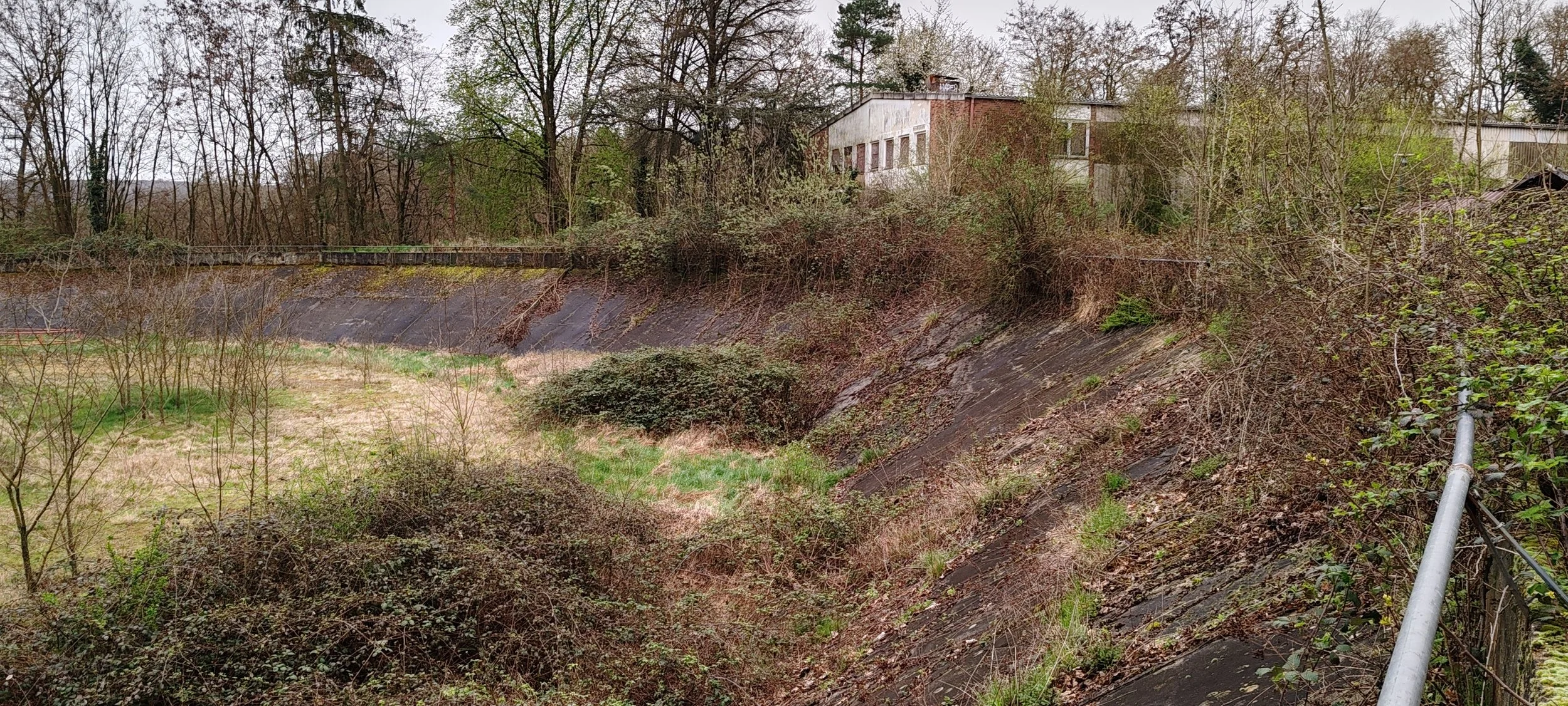 Ein Hang mit Büschen und Bäumen, darunter eine Mauer und ein Gebäude im Hintergrund, an einem bewölkten Tag.