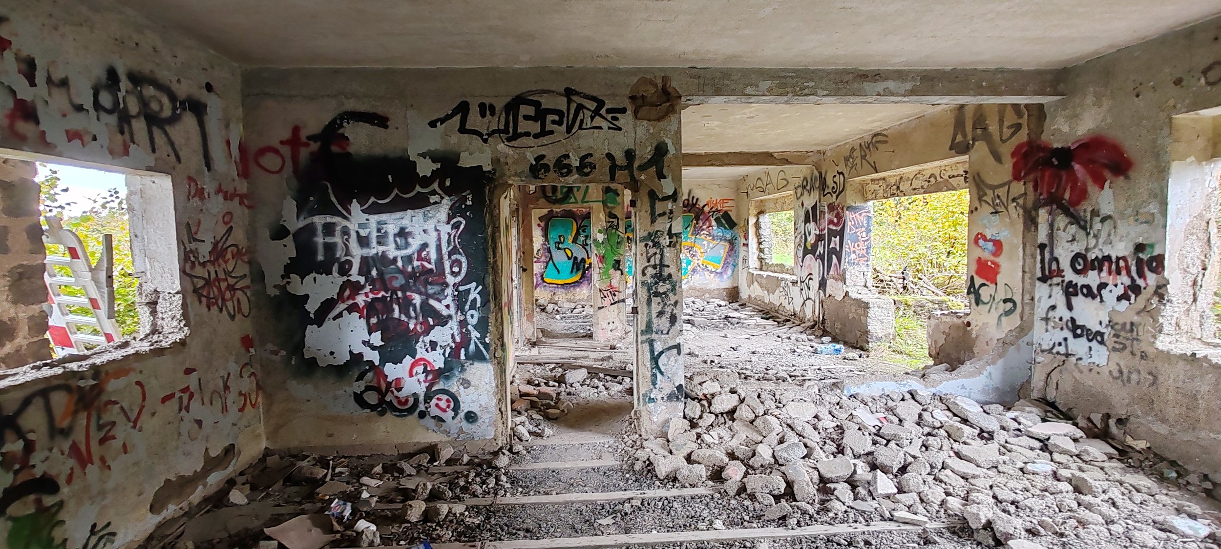 Ein verlassener, zerfallener Bau mit bemalter, mit Graffiti bedeckter Innenwand, durch die Löcher in der Wand und Fenster Blick nach draußen auf Bäume und den Himmel