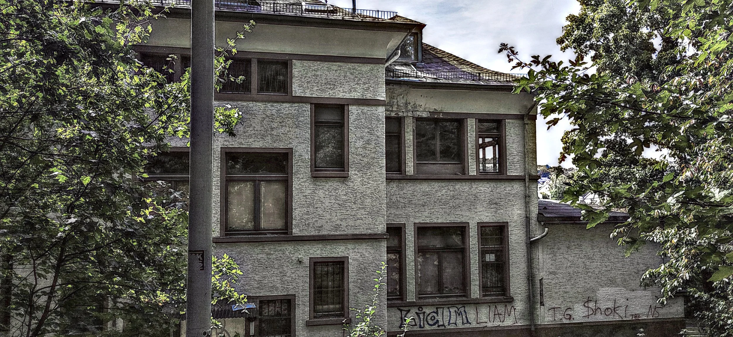 Mehrstöckiges Haus mit grauer Fassade, mehrere Fenster, umgeben von Bäumen, Graffiti an der Wand, städtische Umgebung.