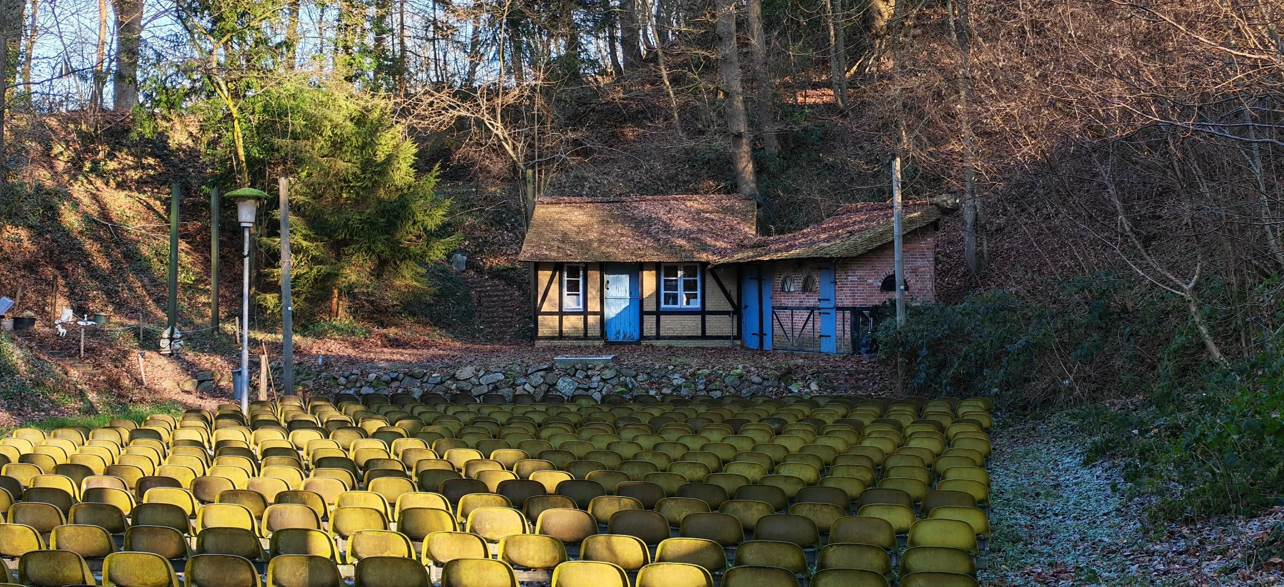 Ein Freilichttheater im Wald mit vielen gelben Sitzreihen vor einer kleinen, alten hölzernen und gemauerten Bühne.