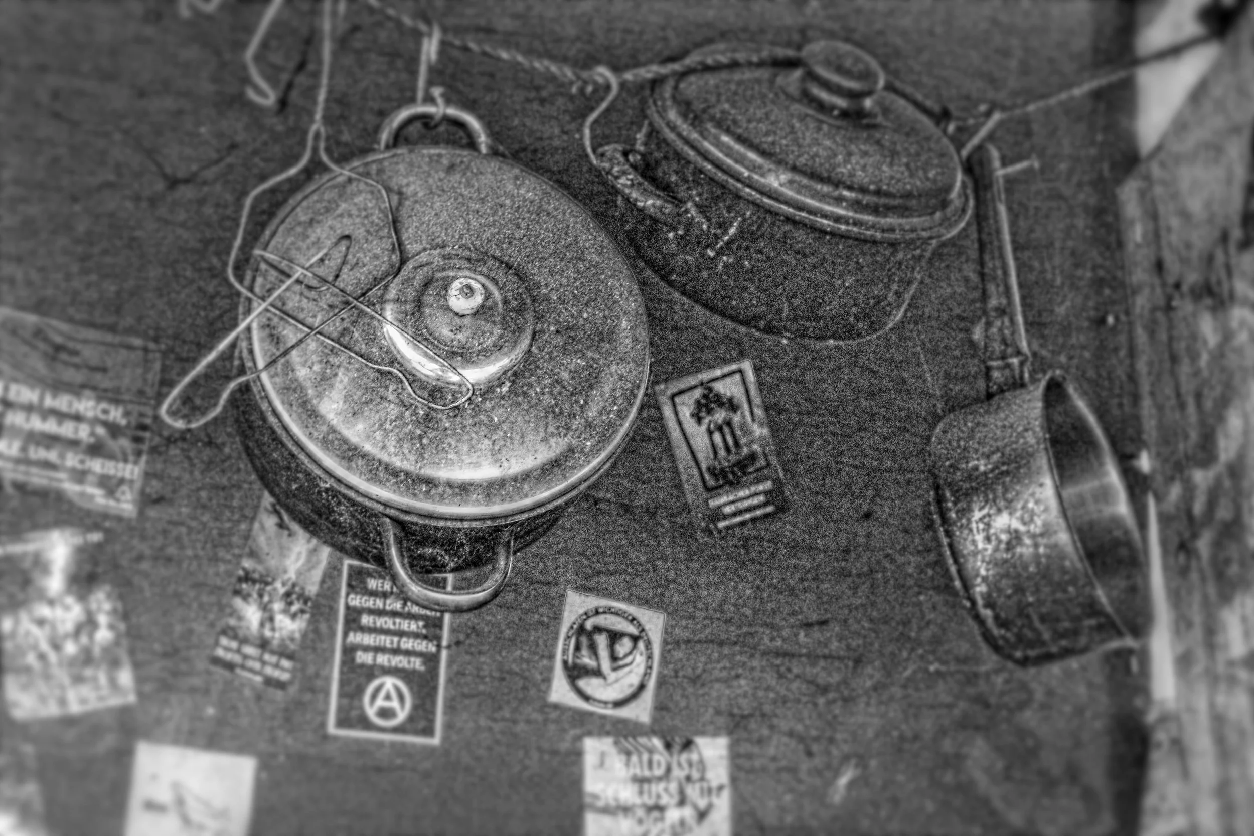 Ein älterer Wasserkocher und eine kleine Metallpfanne auf einer Oberfläche mit mehreren Stickern und Flecken, alles in schwarzer und weißer Farbe.