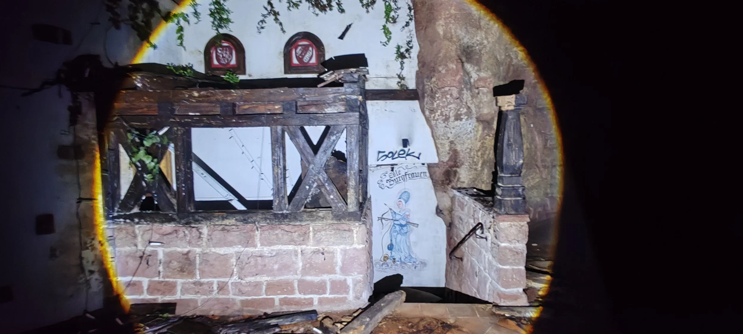Innenansicht mit einer alten Steinkonstruktion, Holzgeländer, Wandgemälde eines Ritters und Graffiti an einer weißen Wand.