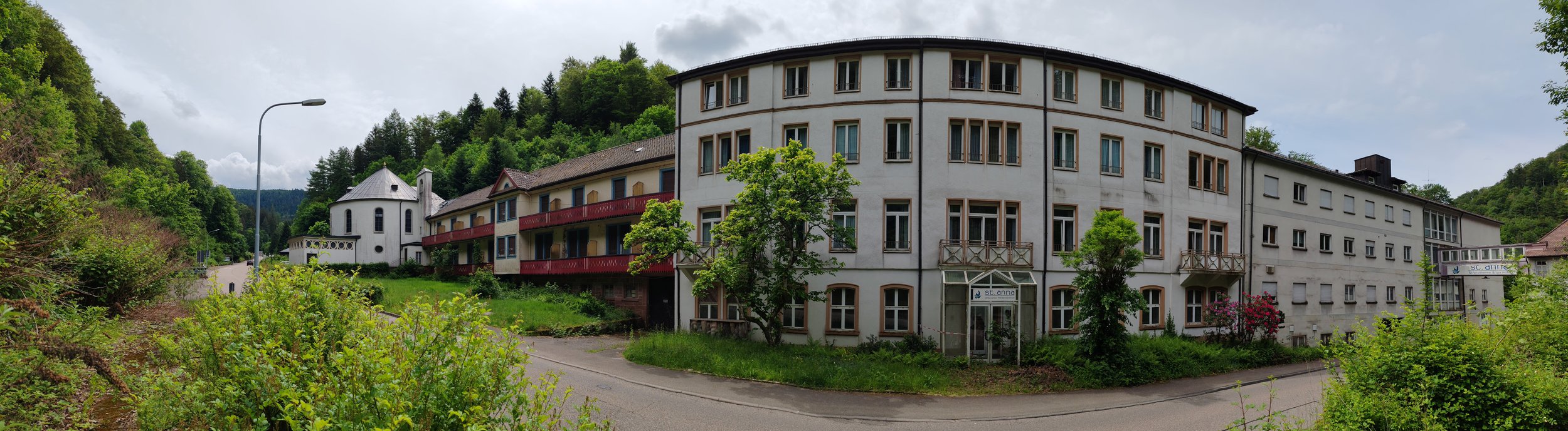 Mehrstöckiges Gebäude inmitten grüner Natur, umgeben von Bäumen und Hügeln, bei bewölktem Himmel.