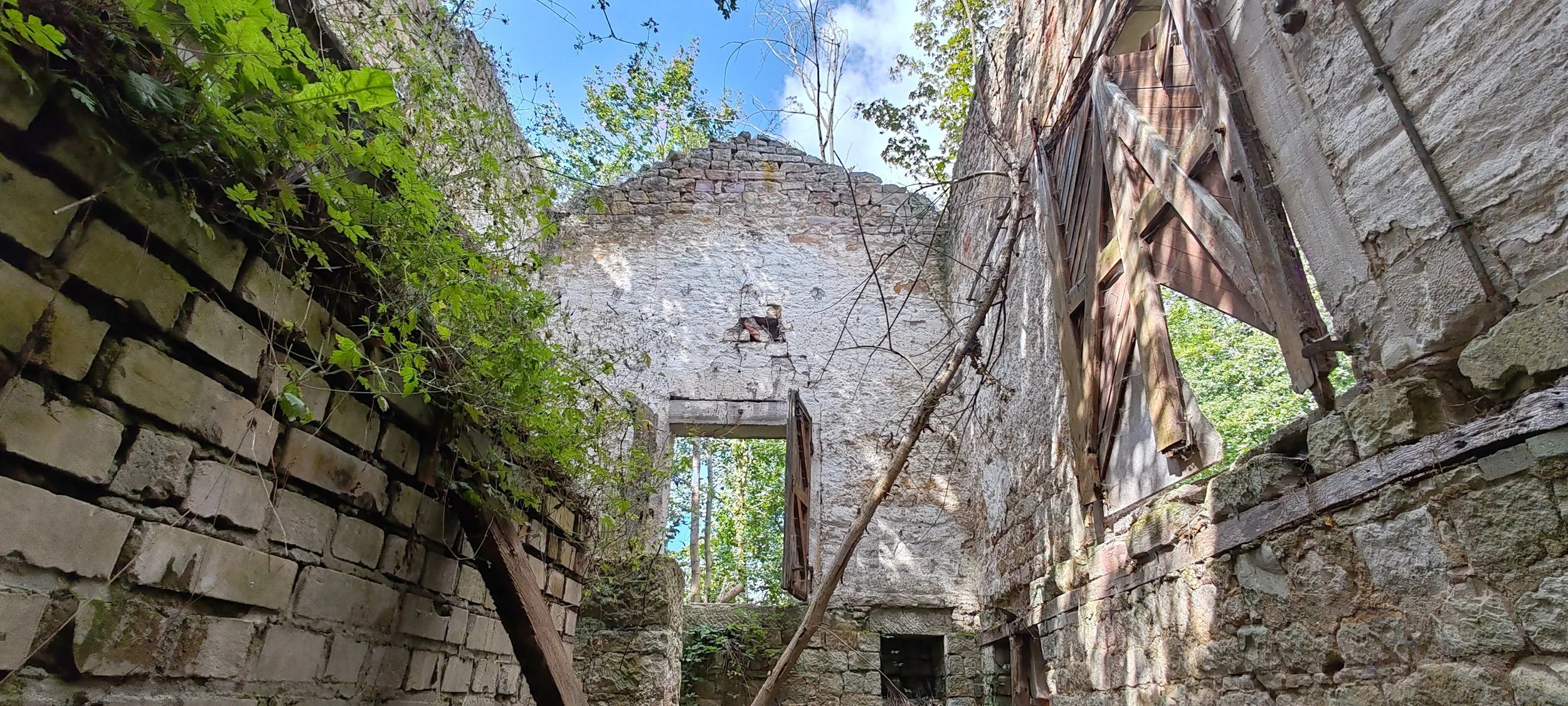 Ruine eines alten Gebäudes mit zerfallenen Wänden, offenen Fenstern mit Holzläden und überhängenden Pflanzen, umgeben von Bäumen unter blauem Himmel.