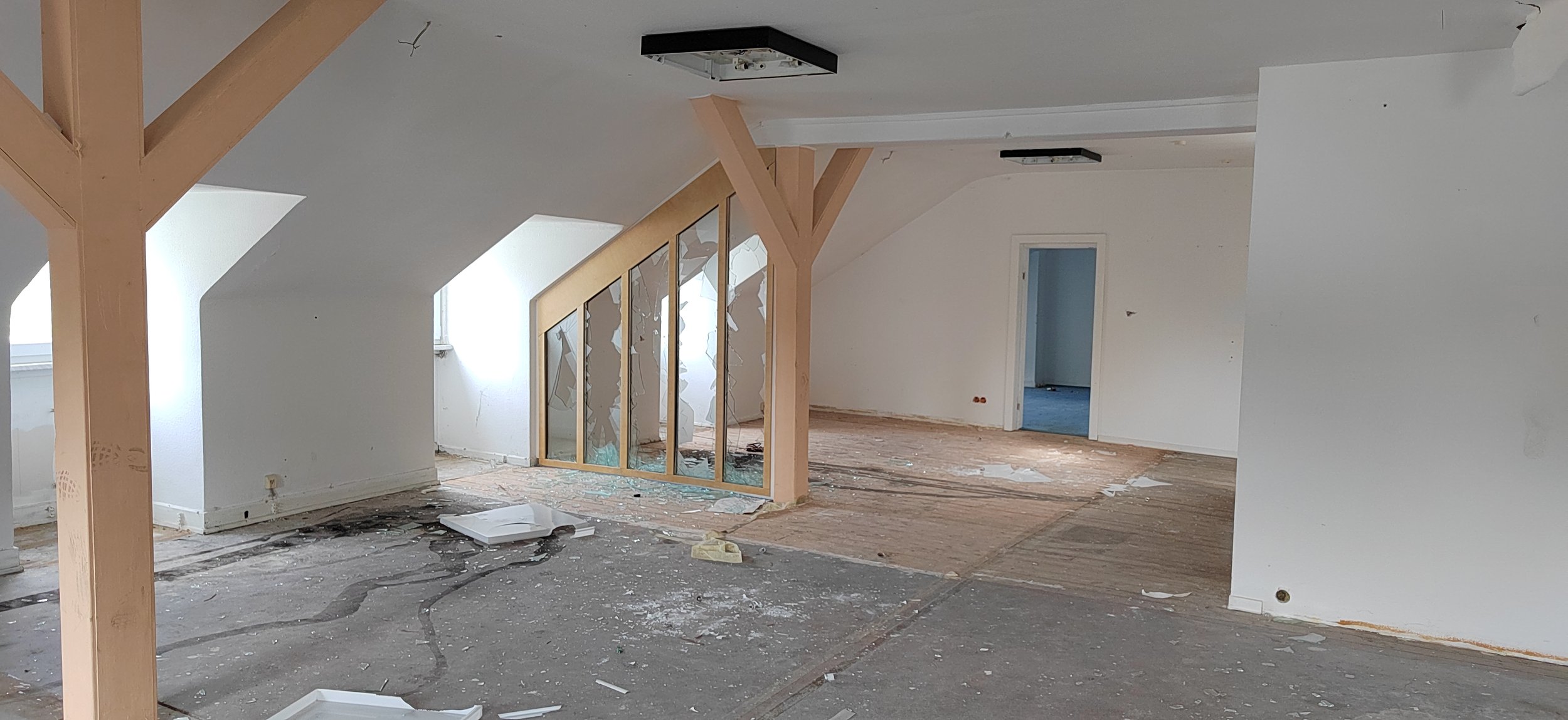 Innenraum eines Hauses, ohne Möbel, mit sichtbaren Schäden an Wänden und Boden, Teile des Fußbodens sind entfernt, Dachbalken sichtbar, einige Fenster und eine Tür sind sichtbar.