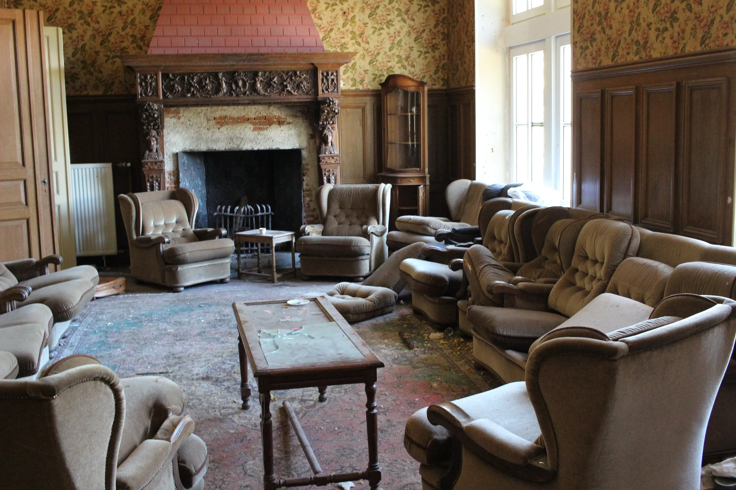Ein alter, verlassener Wohnzimmerraum mit verstaubten, zerknitterten Sesseln, einem mittelgroßen Tisch, einem verzierten Kamin und großen Fenstern, die natürliches Licht hereinlassen, mit abgenutztem Teppich und unordentlichem Zustand.