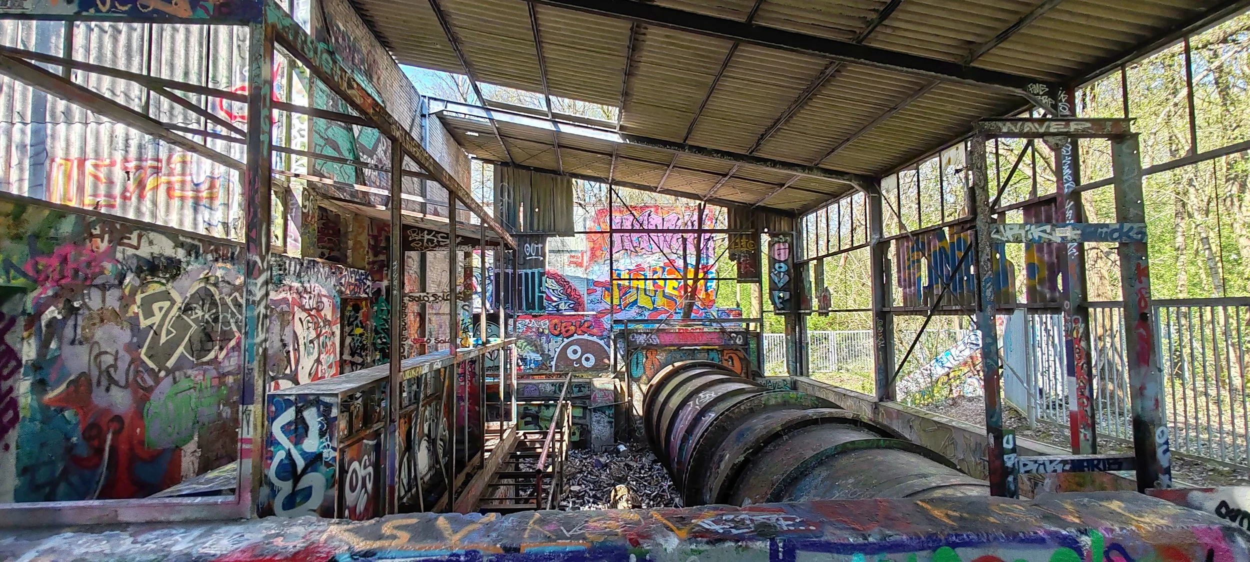Verlassene, mit Graffiti versehene Industriehalle mit Metallkonstruktion, Rutschen und offenen Wänden, umgeben von Bäumen.