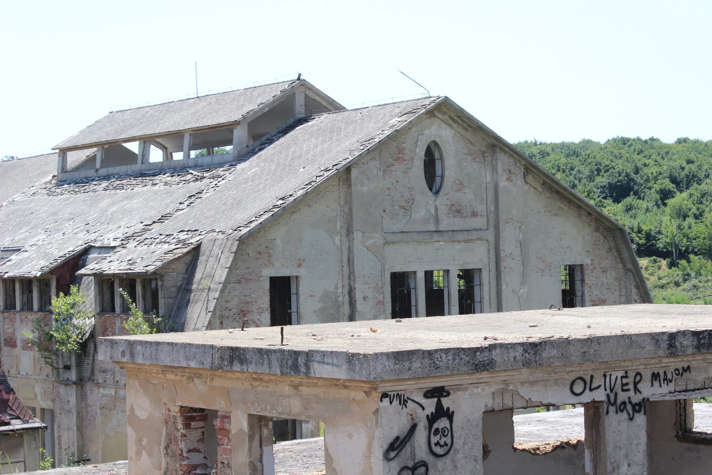 Verfallene, alte Fabrikhalle mit zerbrochenem Dach, grauer Mauer und einem kleinen runden Fenster, im Vordergrund eine betonierte Brüstung mit Graffiti.