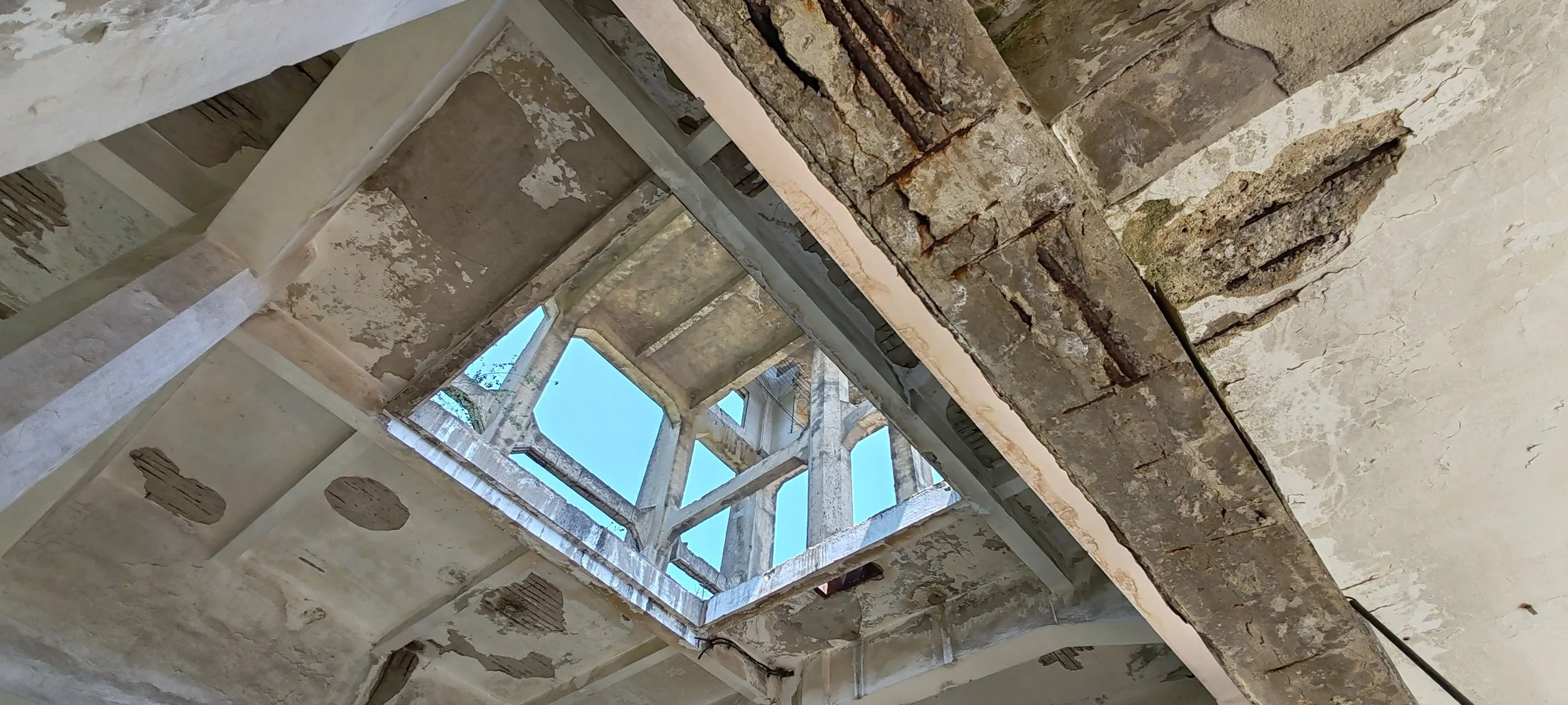 Innenansicht eines alten, verfallenen Gebäudes mit offenen Dachfenstern und sichtbaren, beschädigten Wänden und Decken, Rostspuren auf Metallteilen.