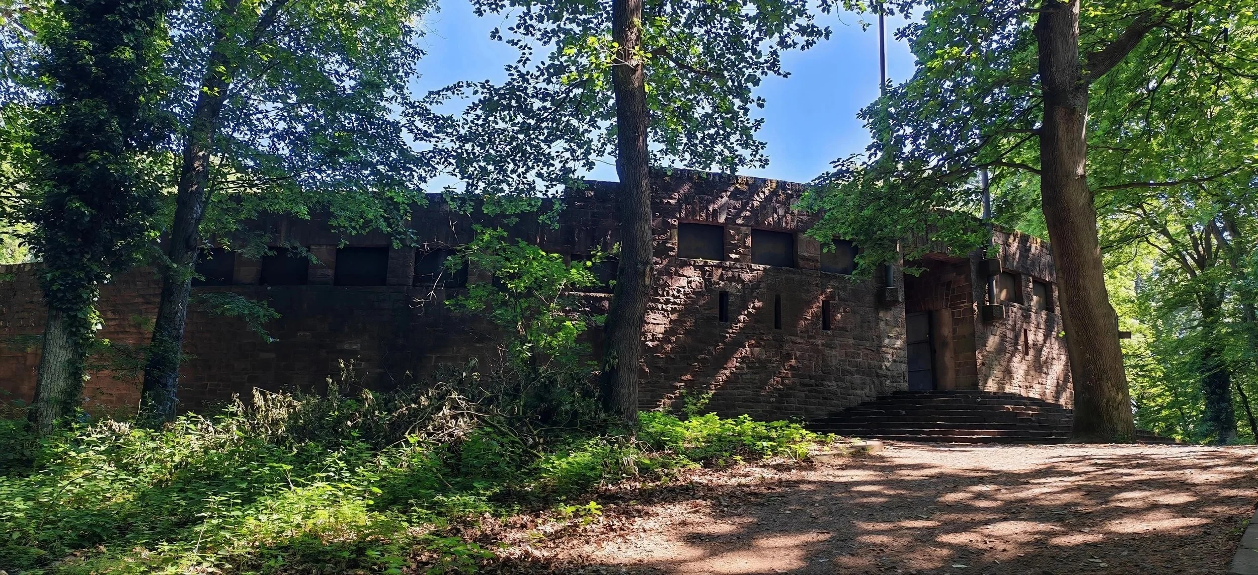 Ein altes Gebäude aus rotem Stein, umgeben von grünen Bäumen im Wald, mit Treppe vor dem Eingang und Sonnenlicht, das durch die Blätter scheint.