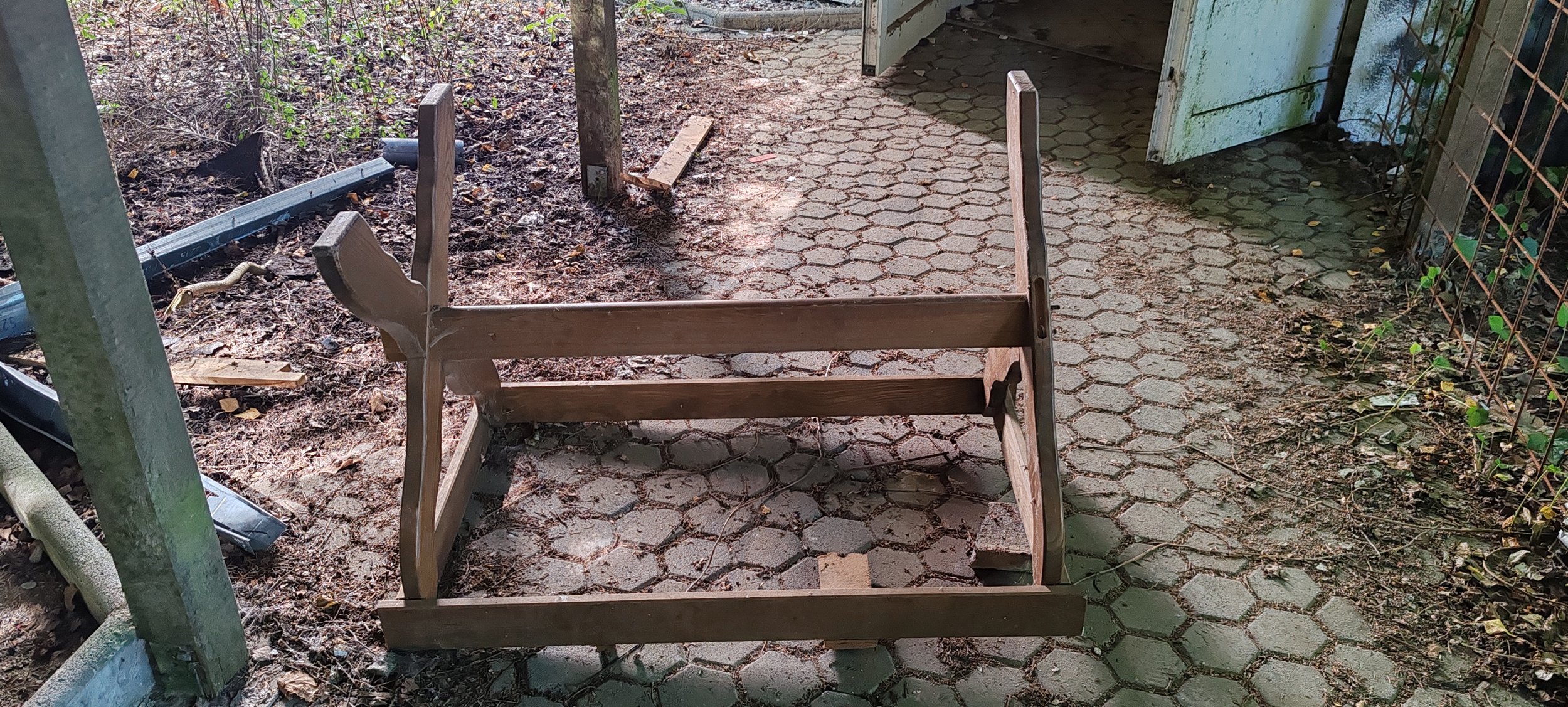 Un abgebautes Holzgestell liegt auf einem harten Boden, umgeben von Staub, Blättern und Werkzeugen, neben einem Gehweg mit hexagonalen Fliesen und einem Zaun aus Metalstäben auf der rechten Seite.