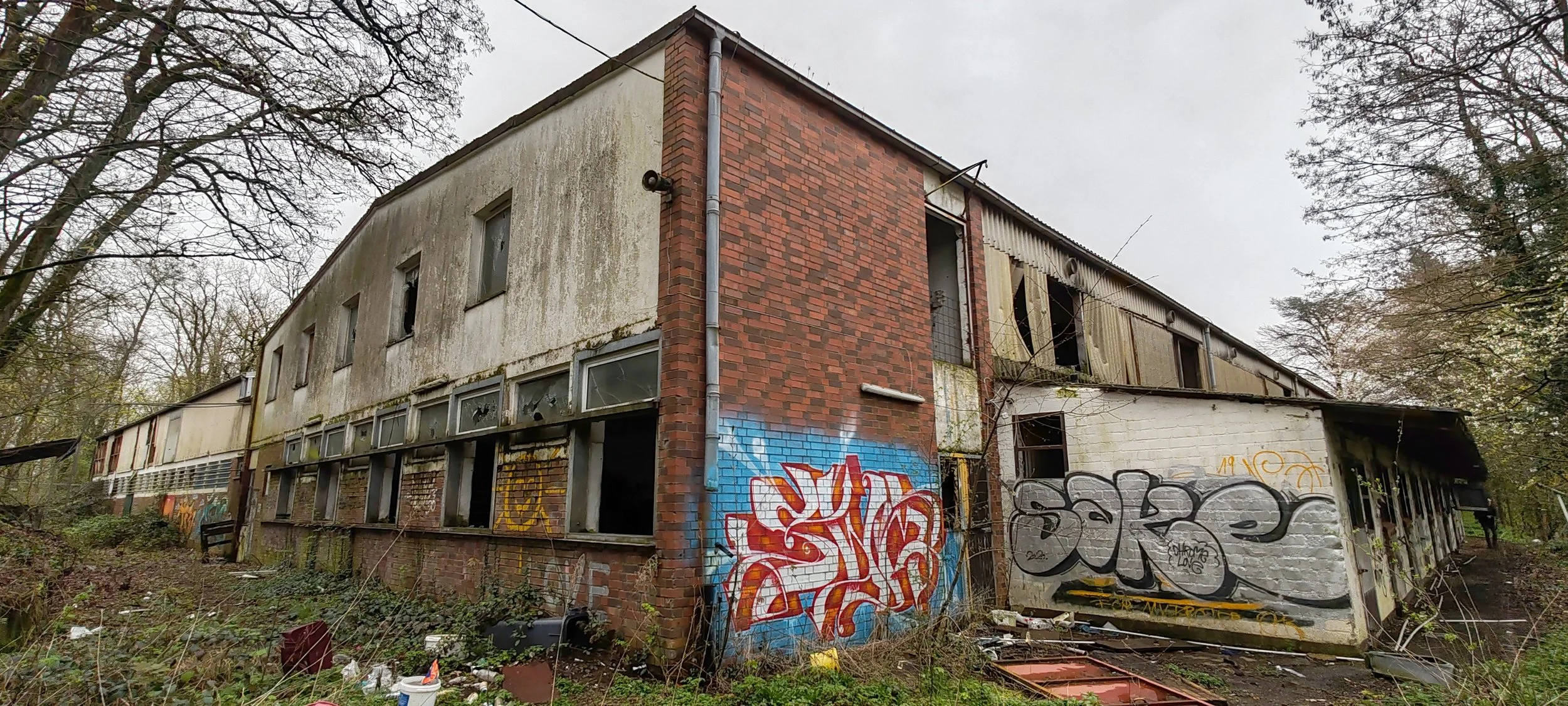 Verwittertes, verfallenes Gebäude mit Graffiti an einer Wand, umgeben von Bäumen und Müll.