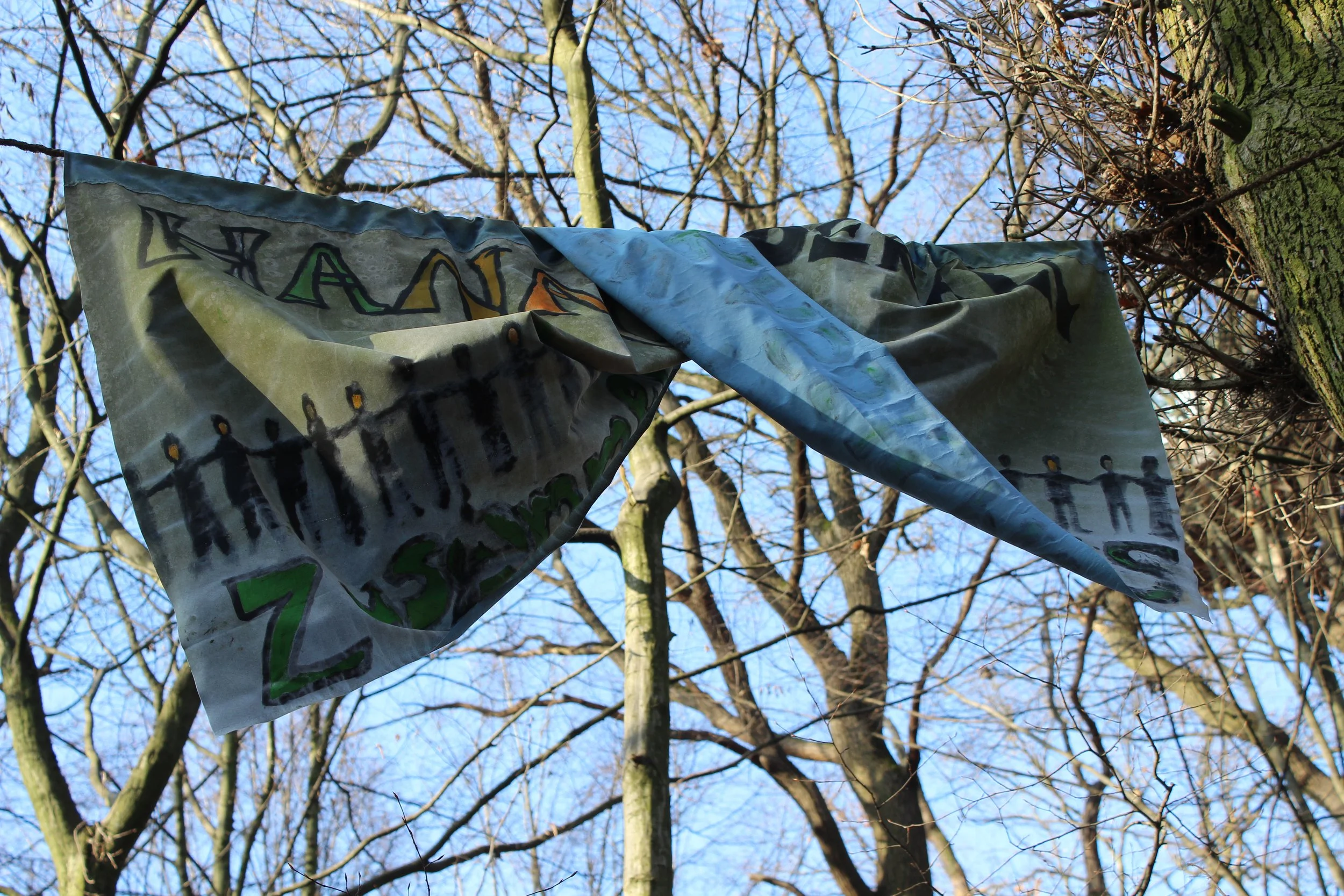 Ein bunt bemalter Stoff hängt an einem Baum im Freien, umgeben von vielen kahlen Baumzweigen und einem blauen Himmel.