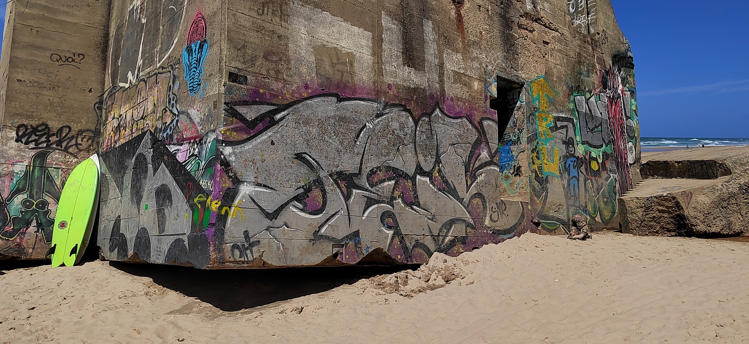 Graffiti an einer Betonmauer an einem Strand mit Sand und sicht auf das Meer, zwei Surfboards stehen an der Mauer, blauer Himmel im Hintergrund.