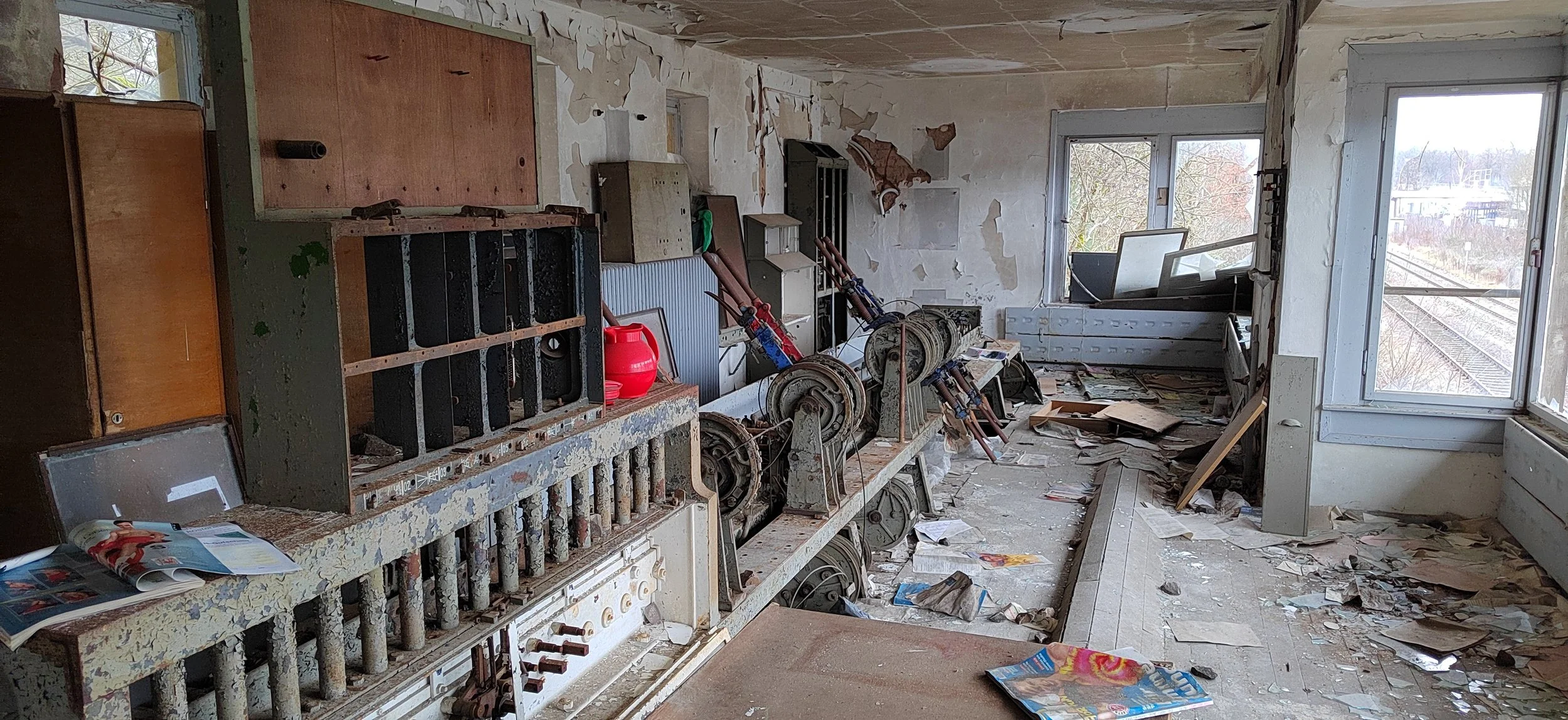 Verlassene, verfallene Innenraum eines Gebäudes mit demolierten Wänden, kaputten Fenstern und unordentlichem Boden mit Papier und Trümmern. Möbel und alte Geräte sind sichtbar.