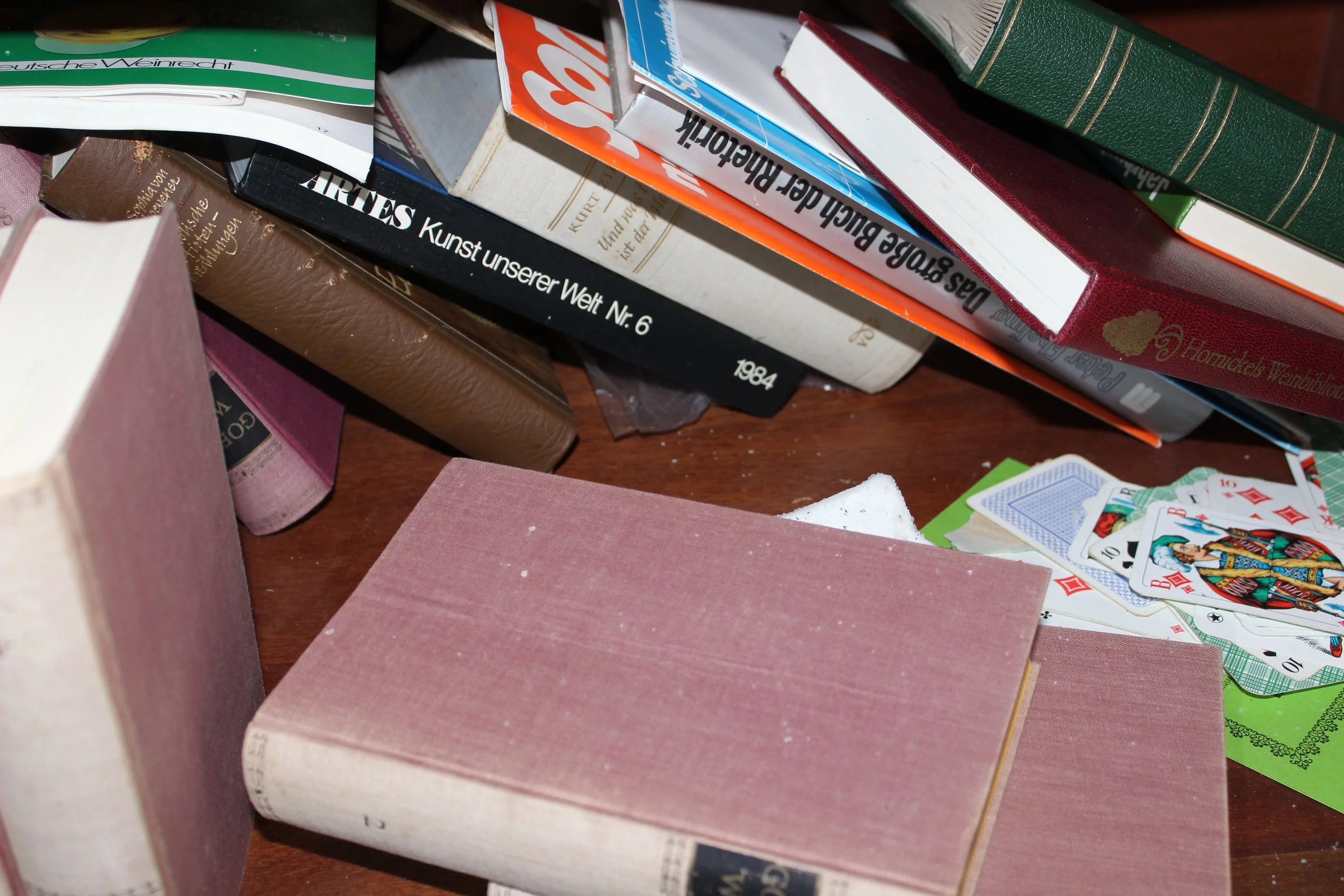 Ein Stapel Bücher und Spielkarten auf einem Holztisch, darunter ein rosa Buchdeckel, die Bücher sind in verschiedenen Farben und Größen. Einige Bücher liegen schräg und andere stapeln sich.