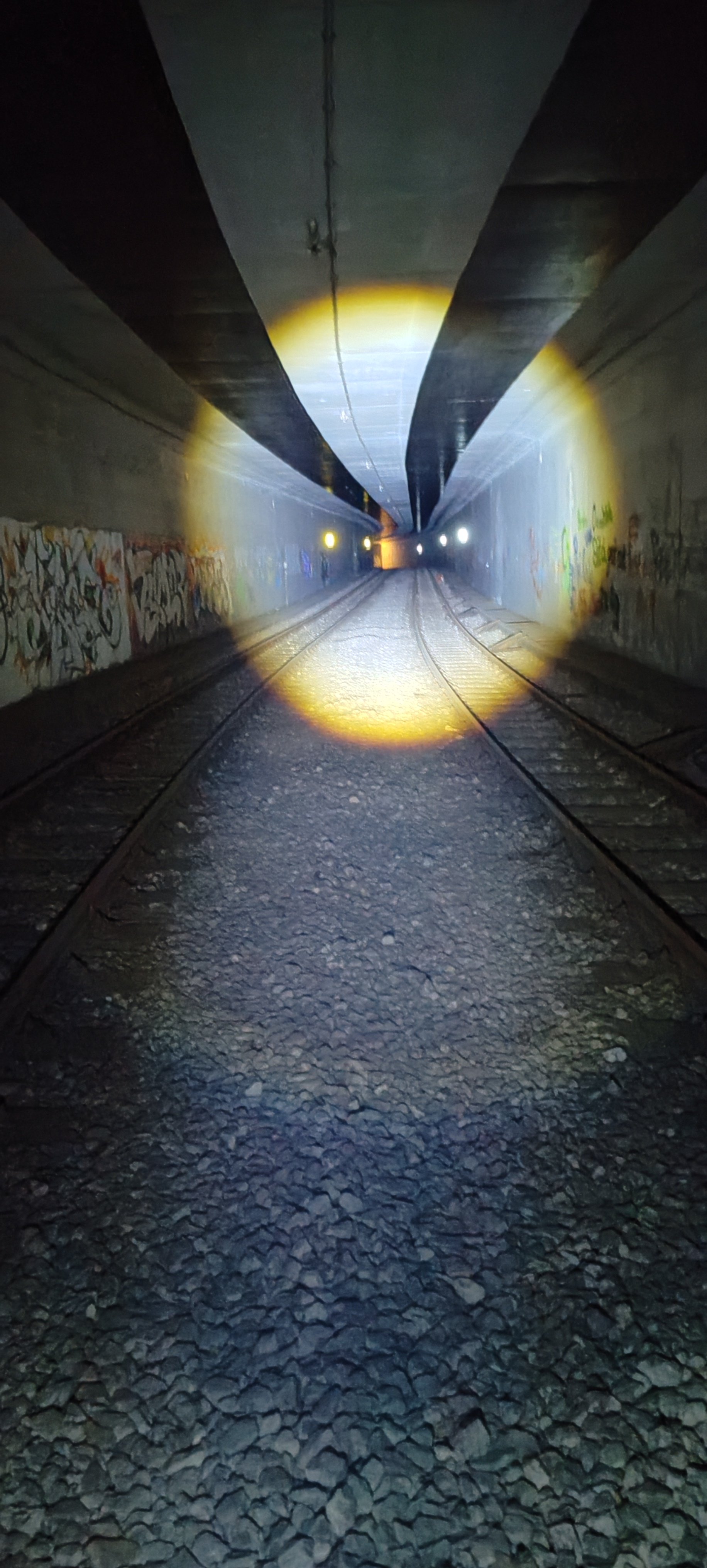 Blick durch eine Tunnel, der mit einem Taschenlampenlicht beleuchtet wird, mit Graffiti an den Wänden und Schienen auf dem Boden.