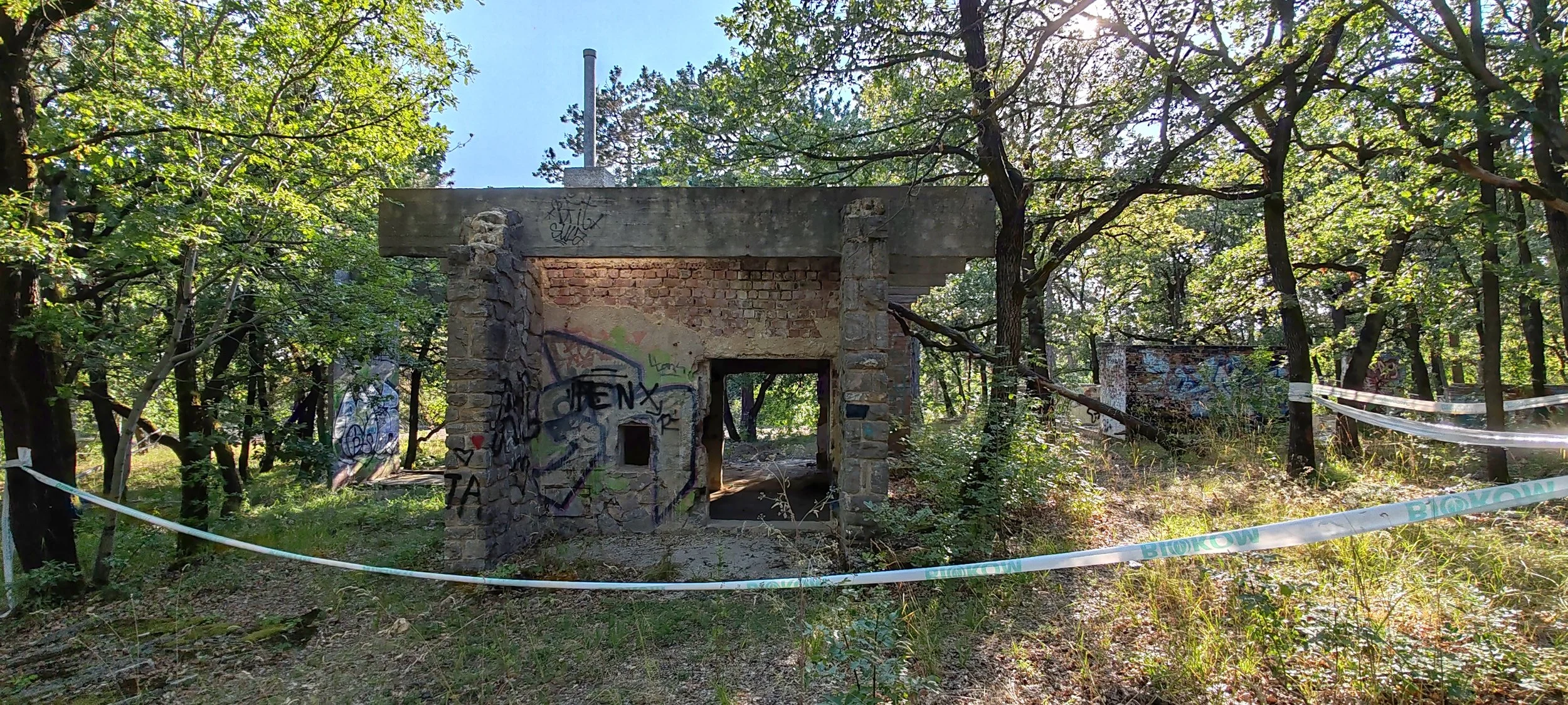 Verfallene, graffiti-bedeckte Gebäude im Wald, umgeben von Bäumen und Sträuchern, mit Absperrband.“